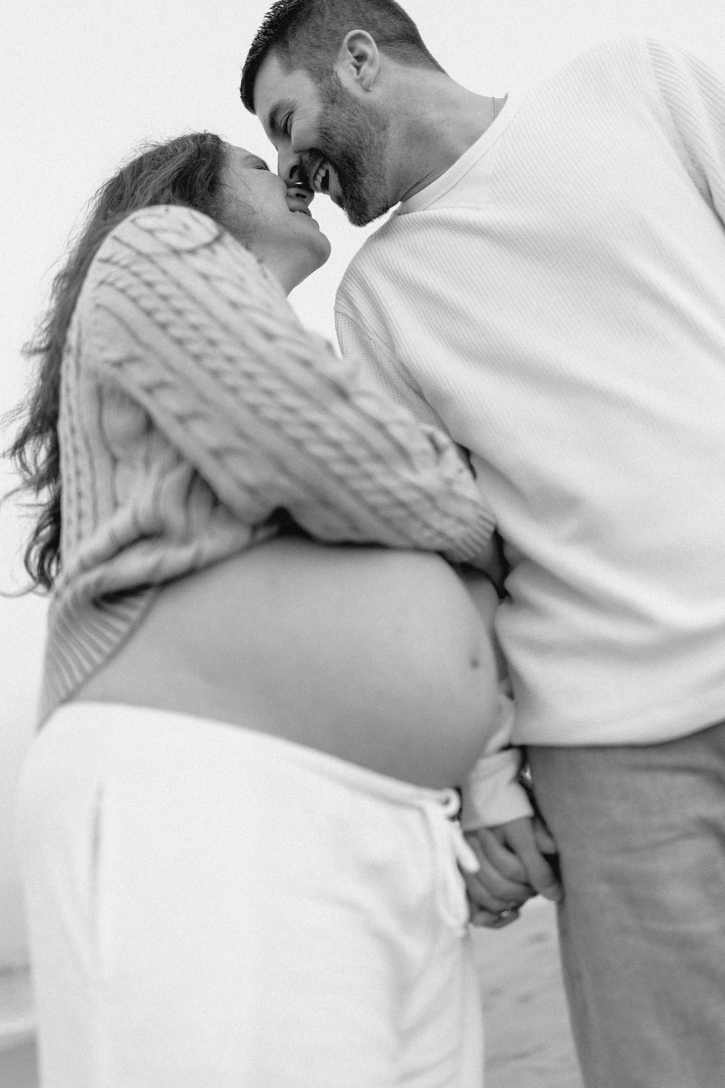 couple laughs and kisses on the beach showing off pregnant belly