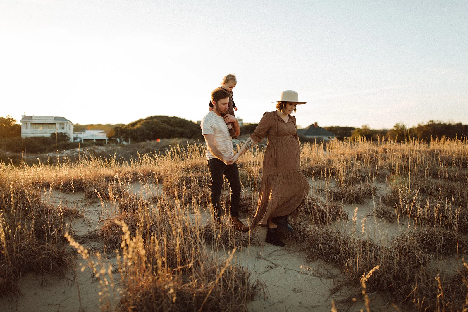 A couple walking hand in hand through a sandy, grassy landscape at sunset, with a young child on the man's shoulders, holding the woman's hand, all dressed in casual, bohemian style.