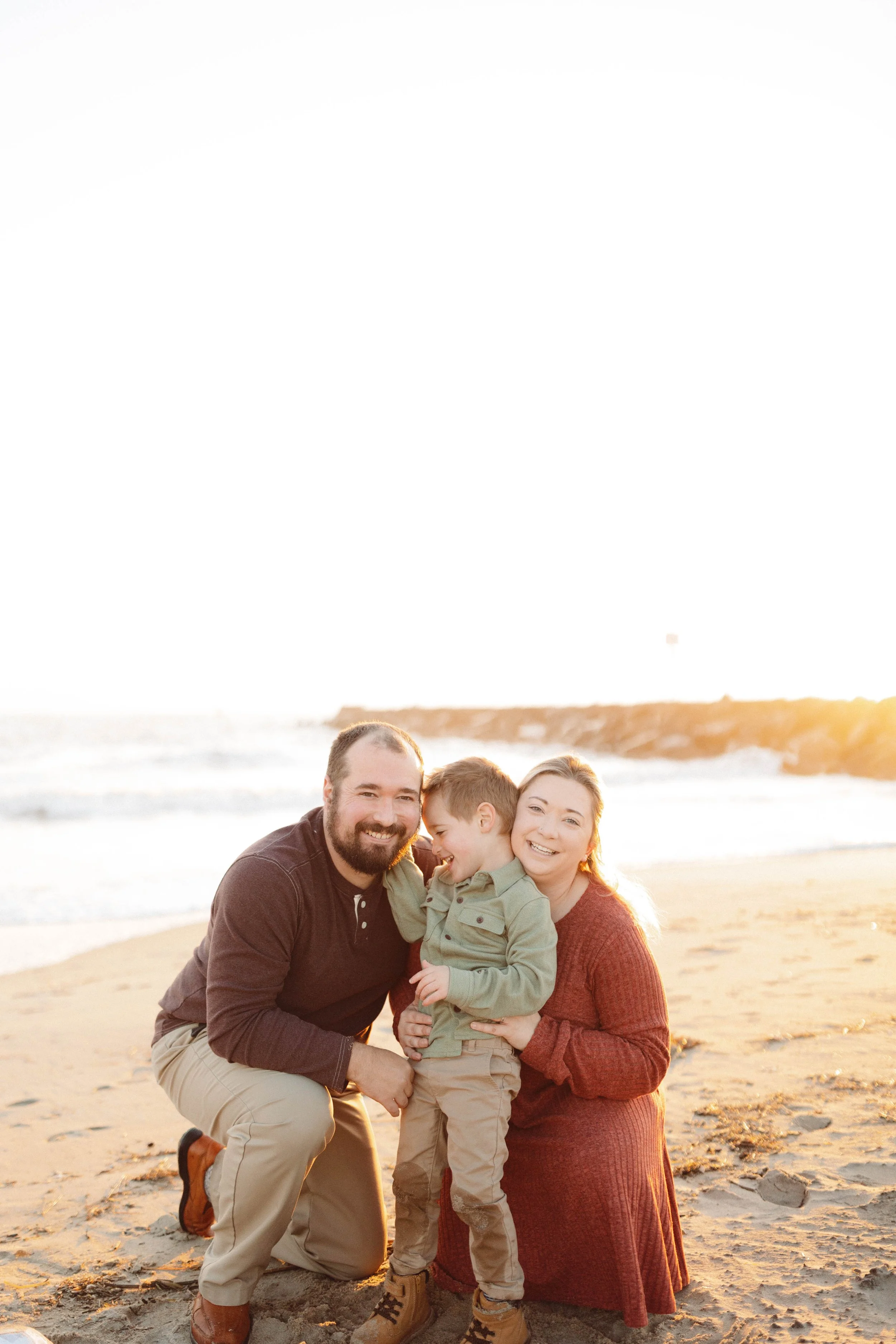 family of three snuggles on a beach while the sun rises over the ocean behind them in virginia beach, virginia