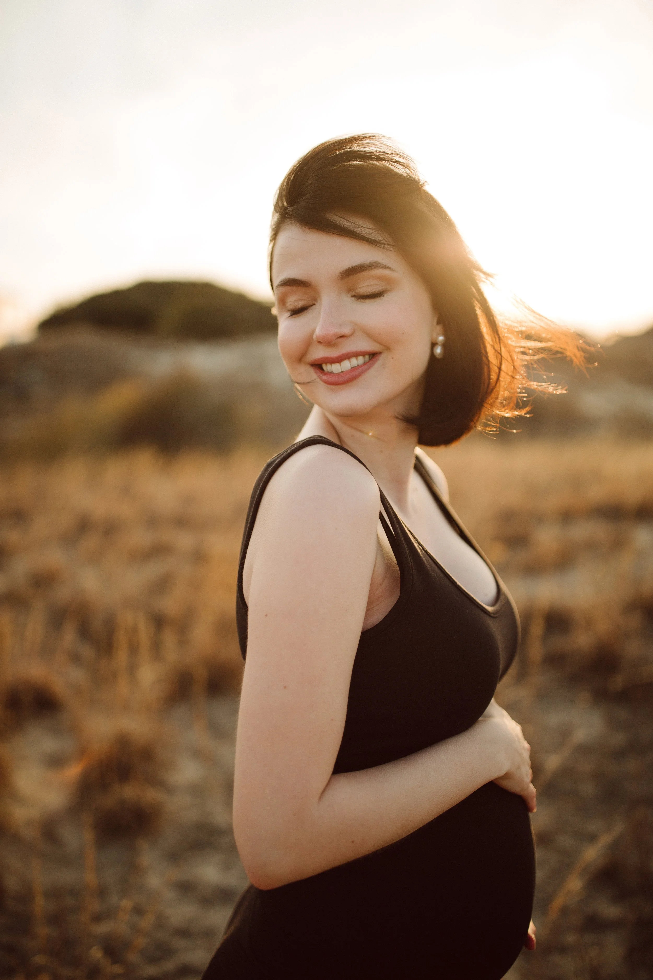 A pregnant woman wearing a black sleeveless top standing outdoors at sunset with a blurred natural landscape in the background, smiling with eyes closed.
