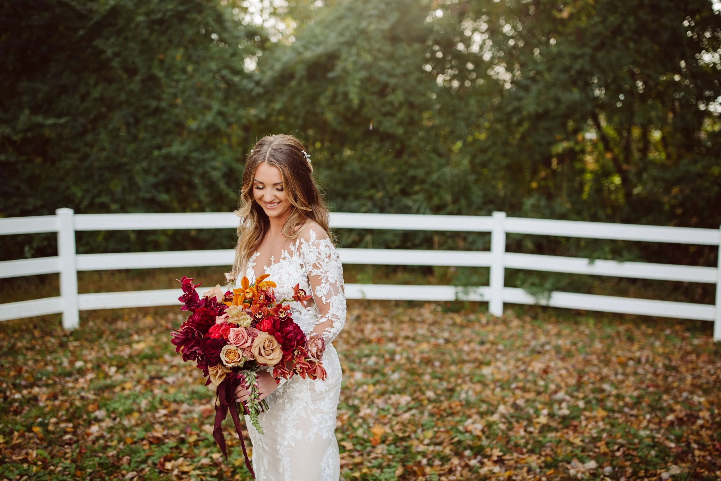 Smiling bride in a white lace wedding dress holding a large bouquet of red, pink, and orange flowers outdoors, with a white wooden fence and trees in the background.