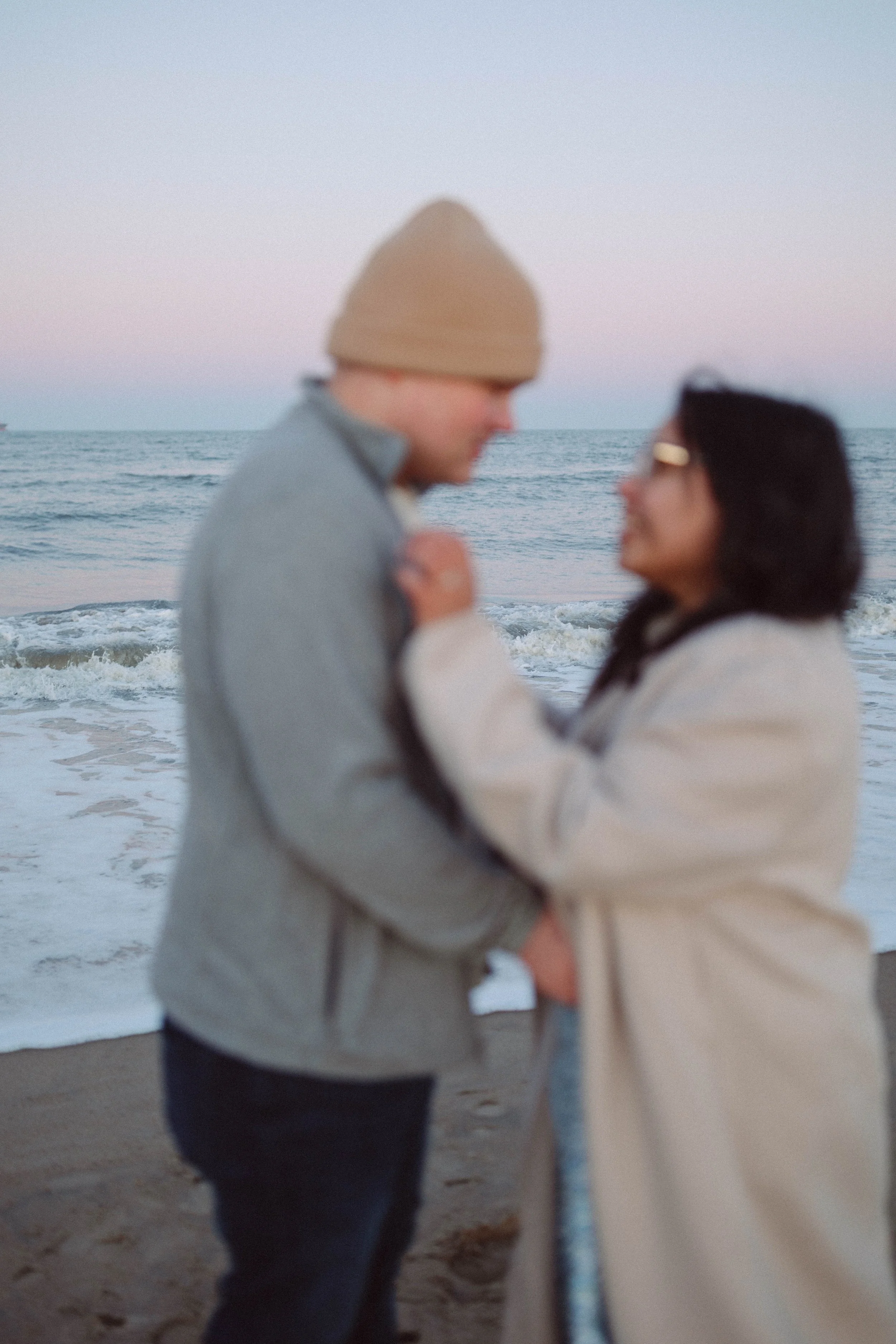 man and woman stand in front of crashing waves on beach looking at each other