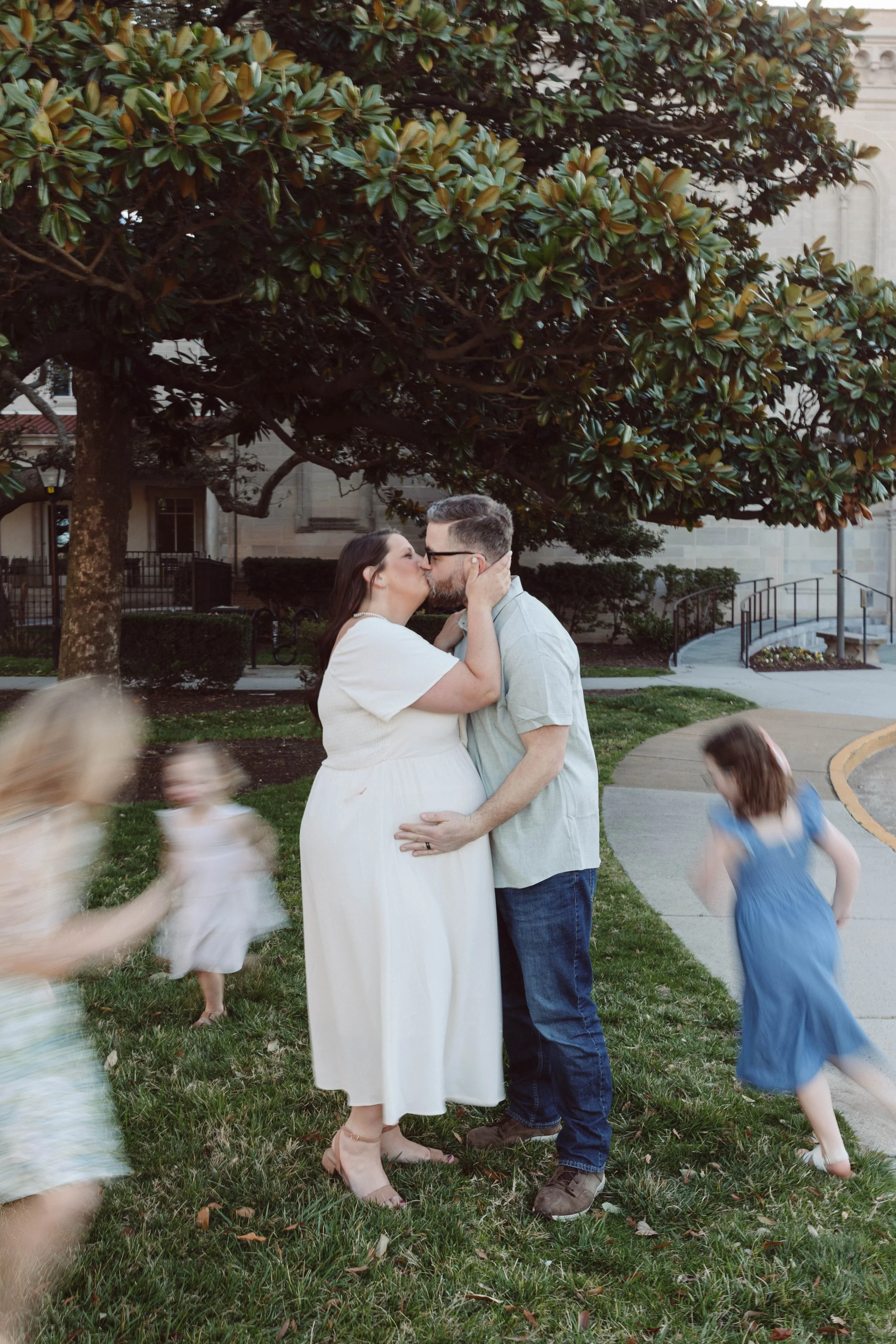 man and woman stand in focus kissing in front of magnolia tree while three children run around them out of focus