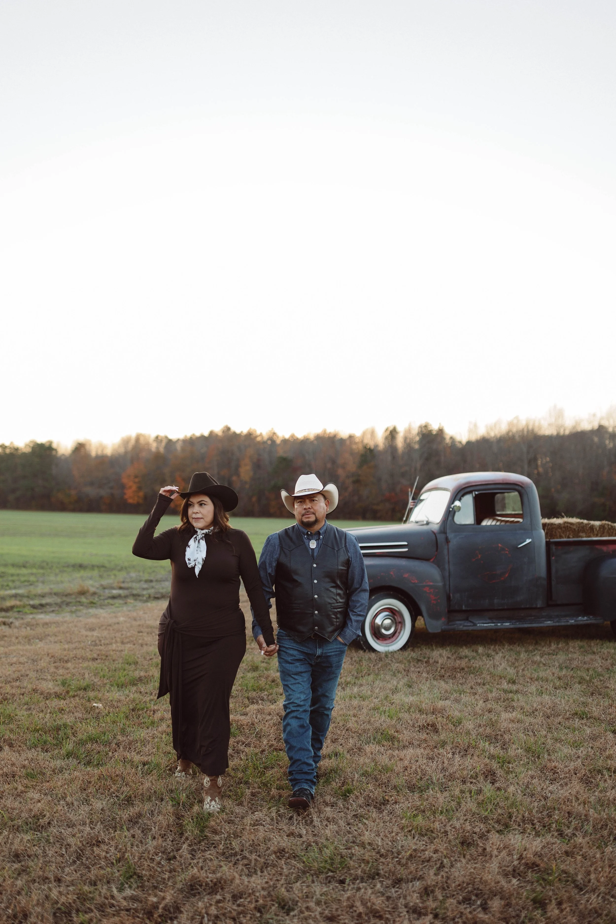 couple in western attire walks through a field looking toward the horizon with an antique ford pickup truck in the background