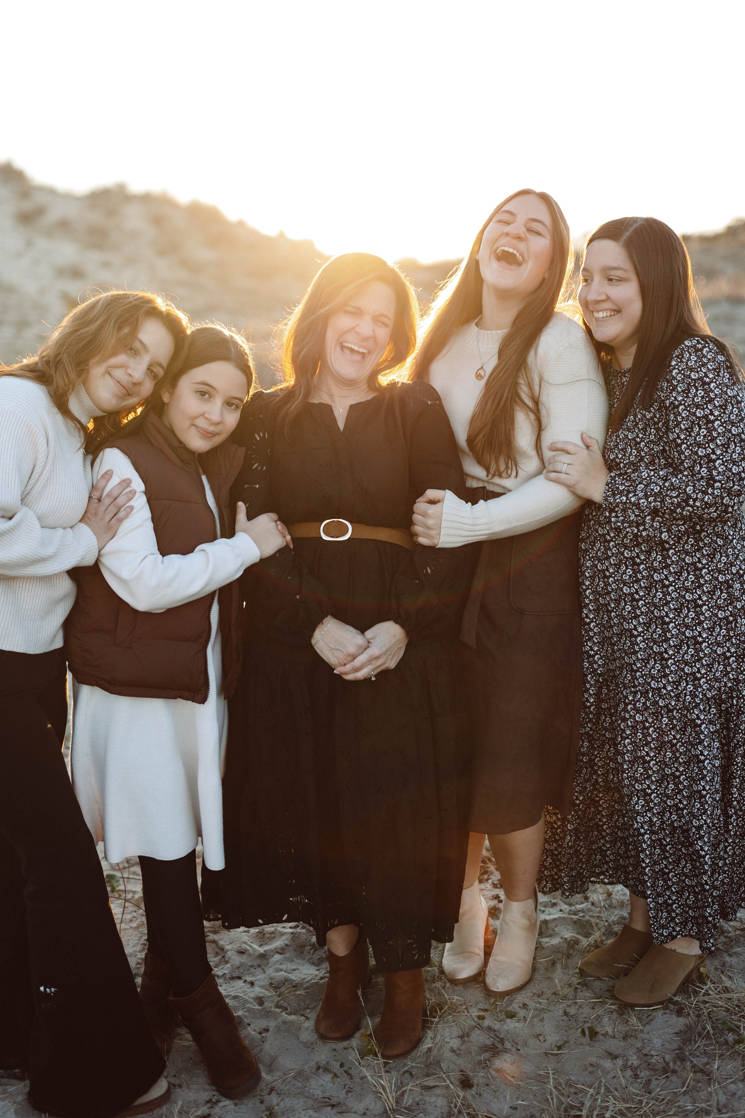 group of women laughing toward the camera while standing on sand dunes with the sun setting behind them, wearing coordinated outfits of tan and black.