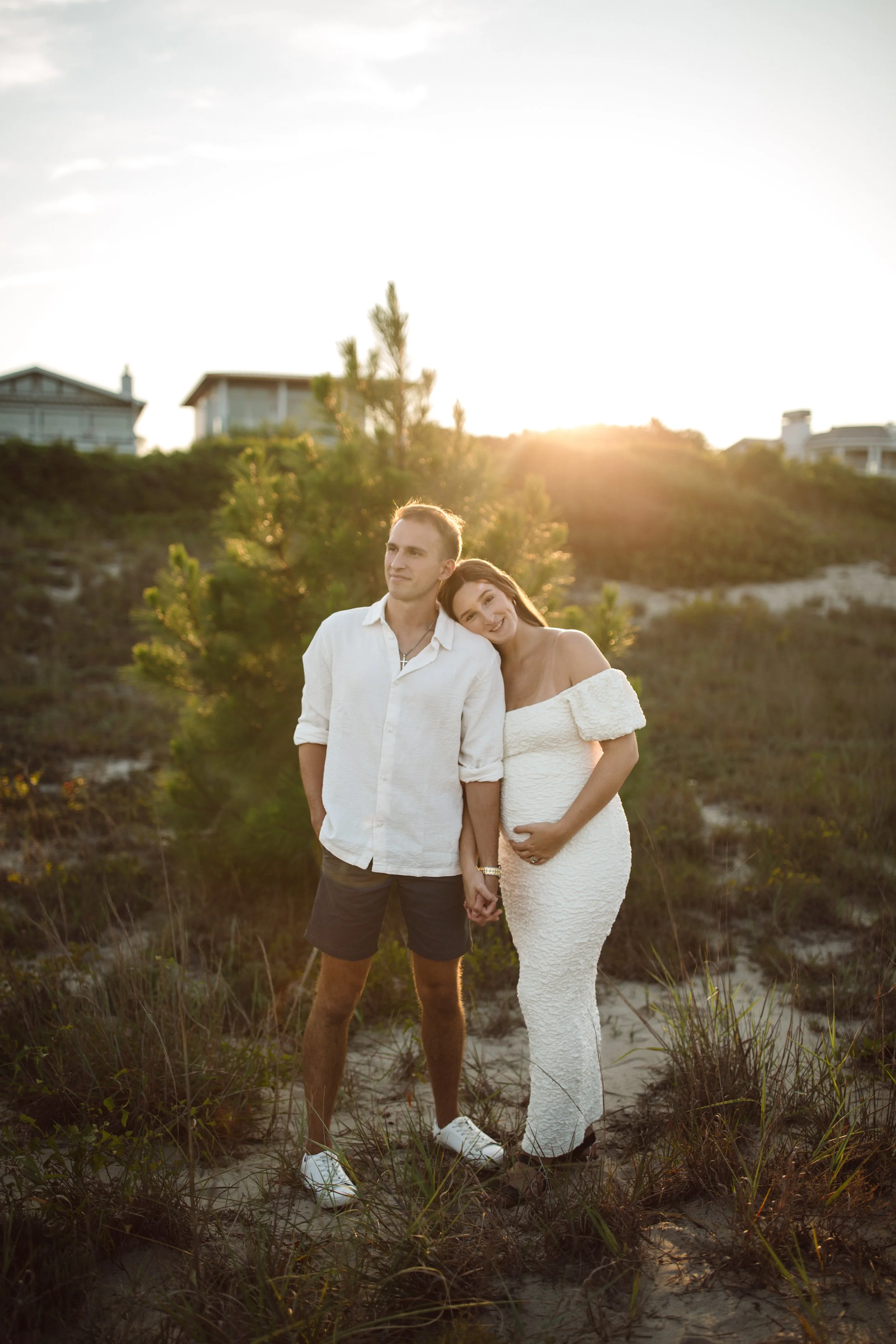 A couple stands on a sandy beach holding hands, with the pregnant woman resting her head on the man's shoulder, during sunset. The woman wears an off-shoulder white dress, and the man wears a white shirt and dark shorts. There are houses on a hill in