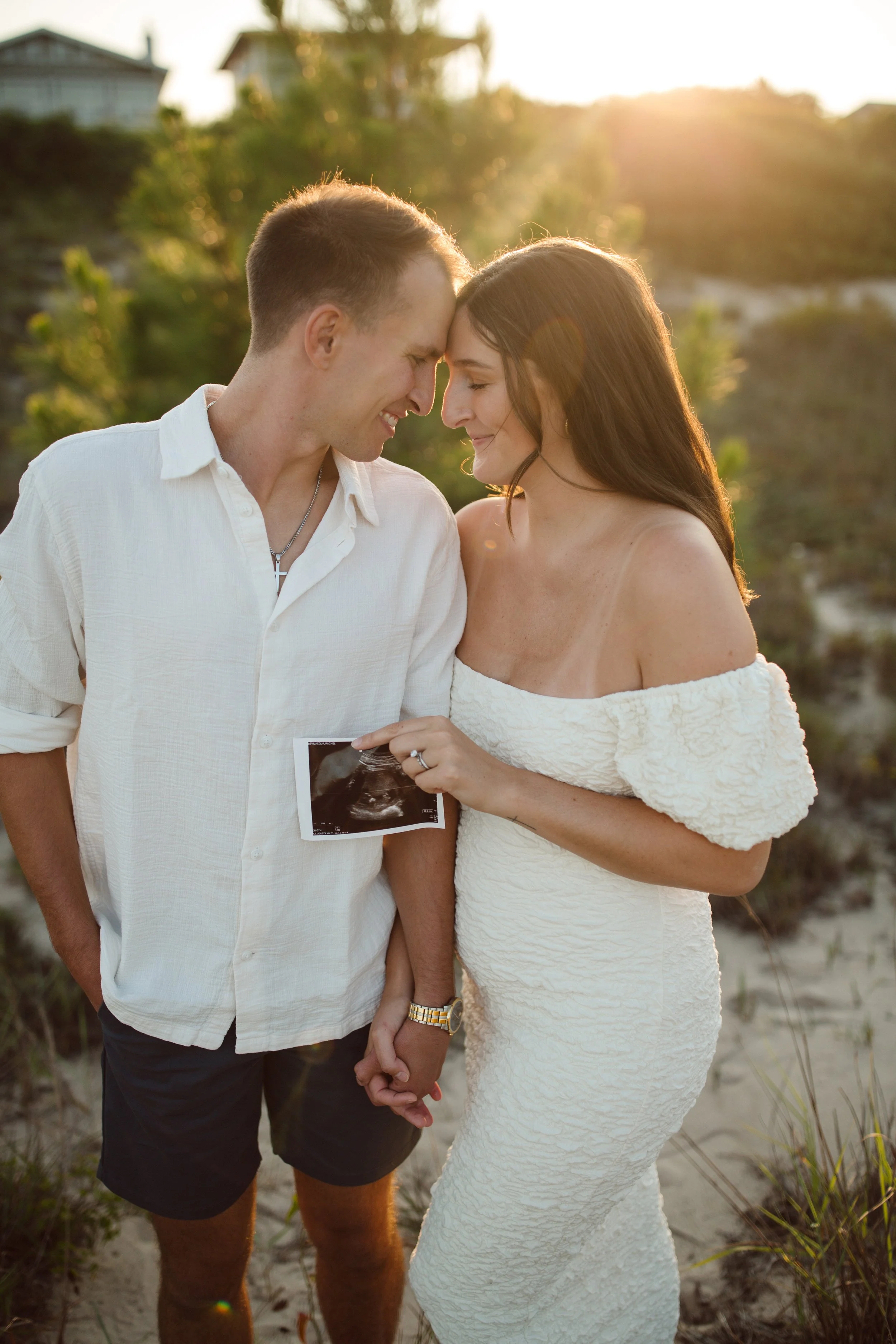 A couple is holding hands and smiling at each other while standing on a beach at sunset. The woman is holding an ultrasound image, indicating pregnancy.
