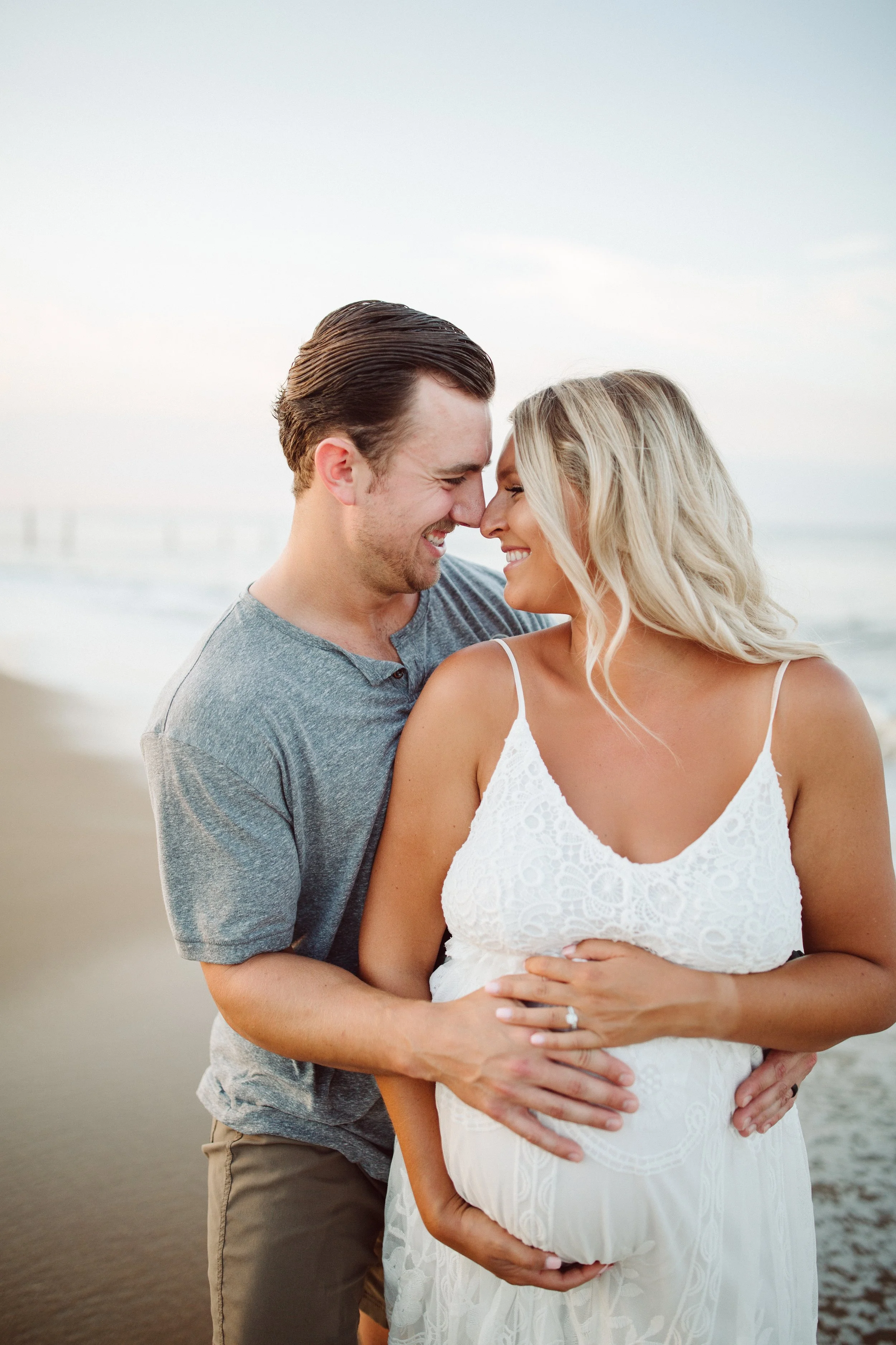 A smiling couple in casual clothing stand close on the beach, with their foreheads touching and hands on a pregnant woman's belly.
