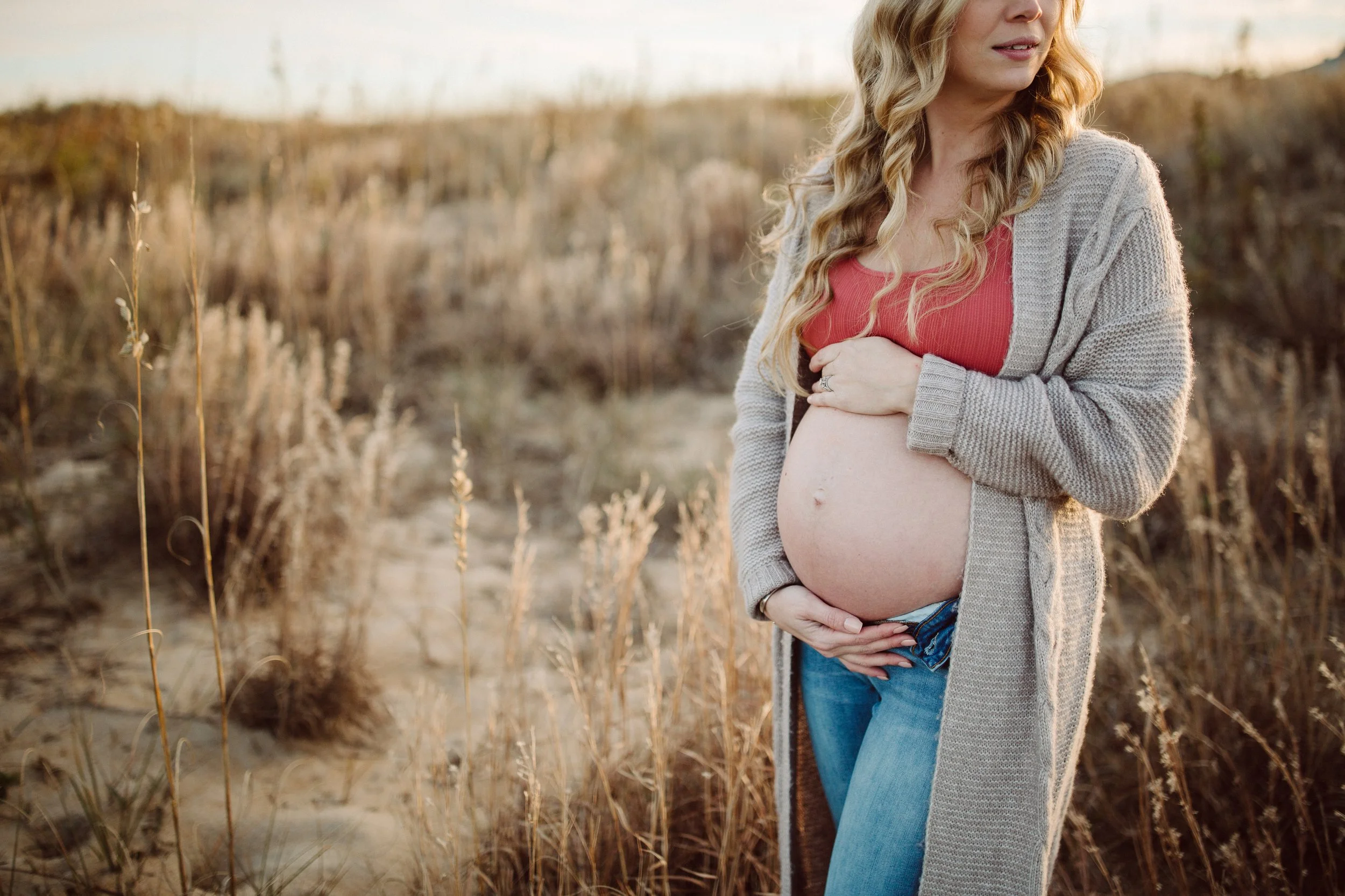 A pregnant woman with long, wavy blonde hair standing in a field of tall, dry grass, wearing a red top, a grey cardigan, and blue jeans, gently holding her belly with both hands. The scene is lit by warm sunlight.