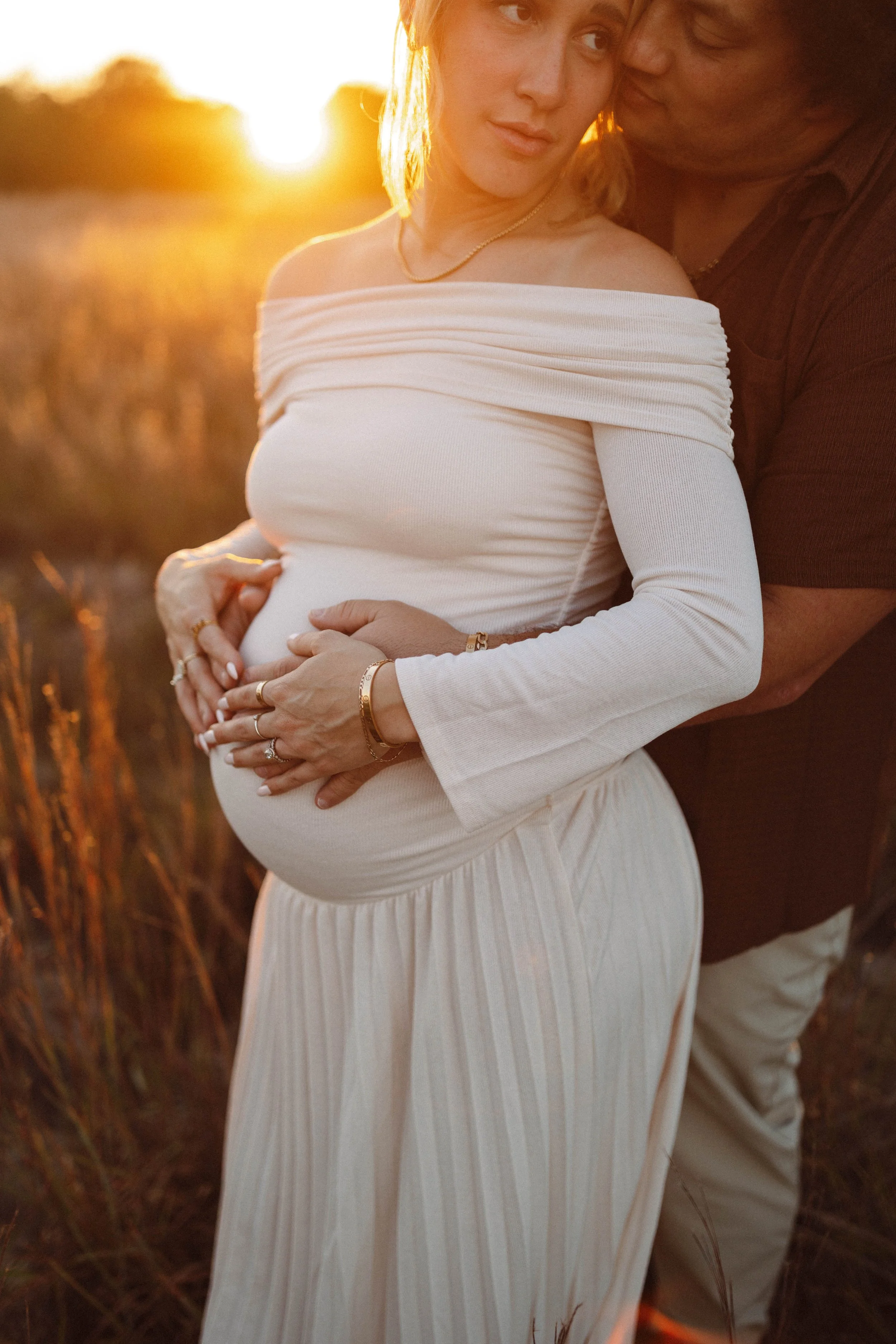 A pregnant woman in a white off-shoulder dress with wavy blonde hair and a man in a dark shirt in a field at sunset, both touching her belly.