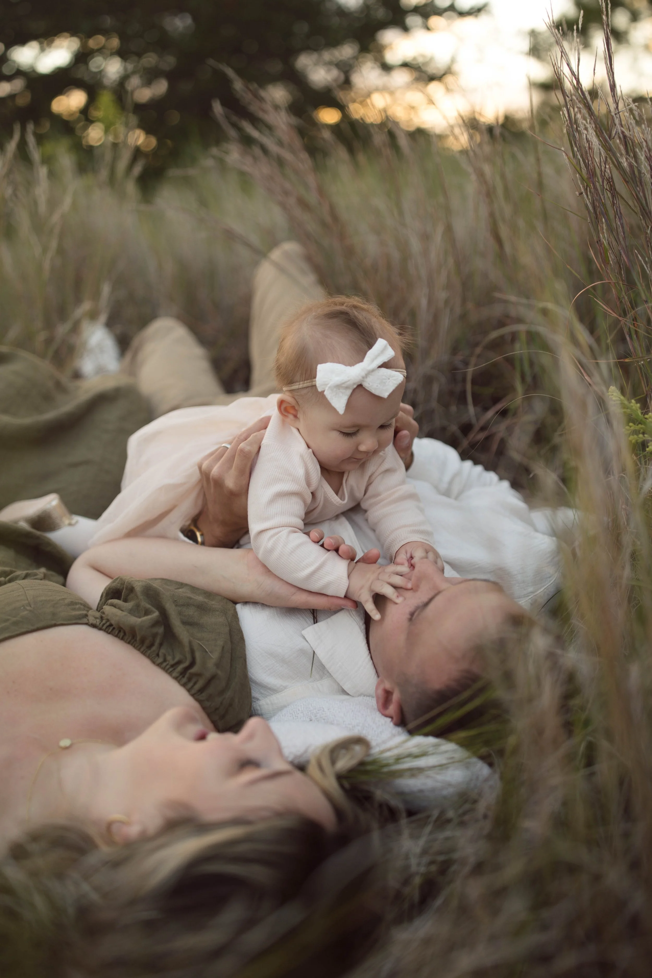 A woman, man, and baby lie on the ground in a grassy field during sunset, playing together and smiling.