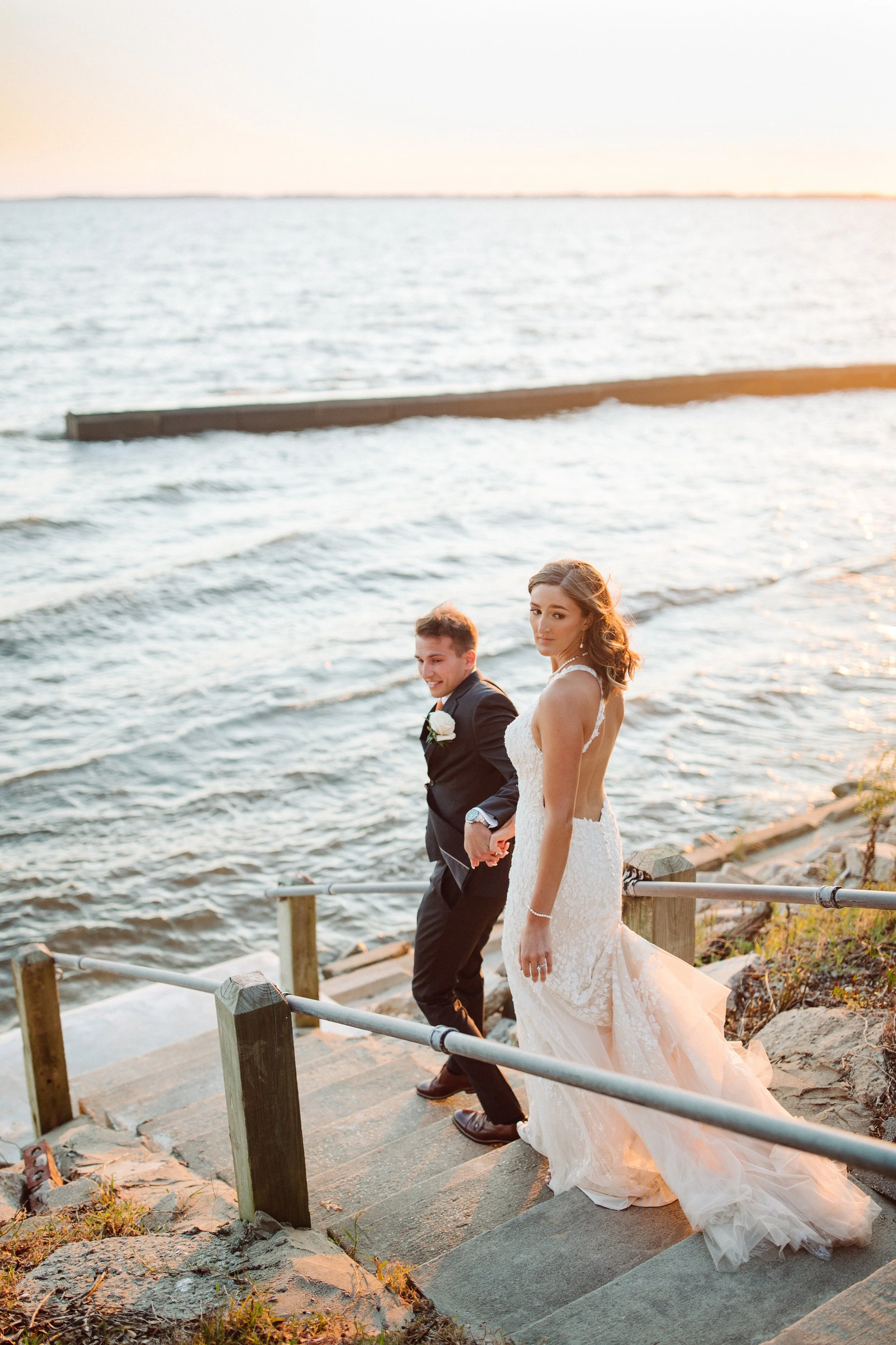 A newlywed couple in wedding attire walking up outdoor steps along a waterfront at sunset.