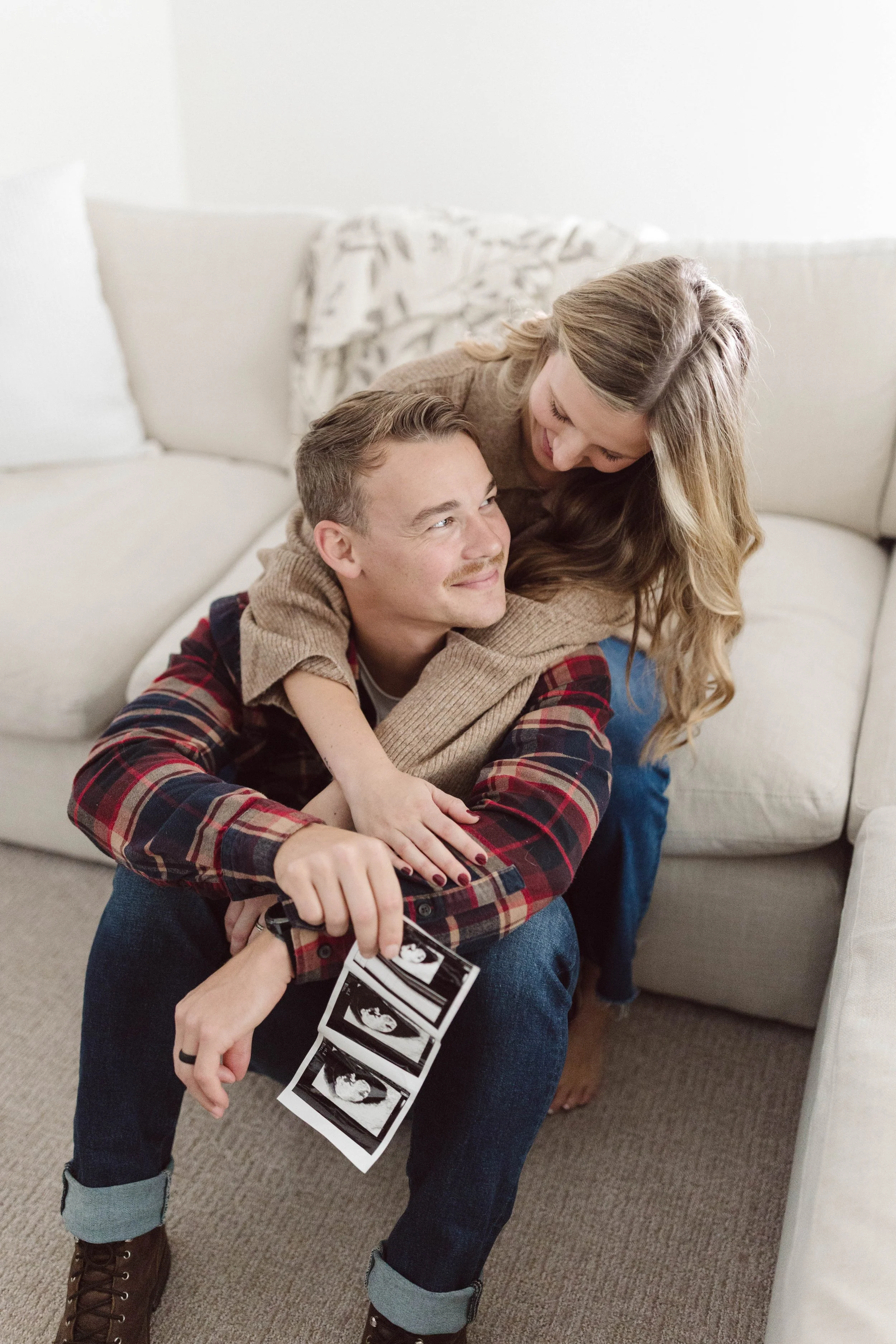 A couple sitting on the floor in a living room, holding ultrasound photos, with the woman hugging the man. They are smiling, celebrating a pregnancy.