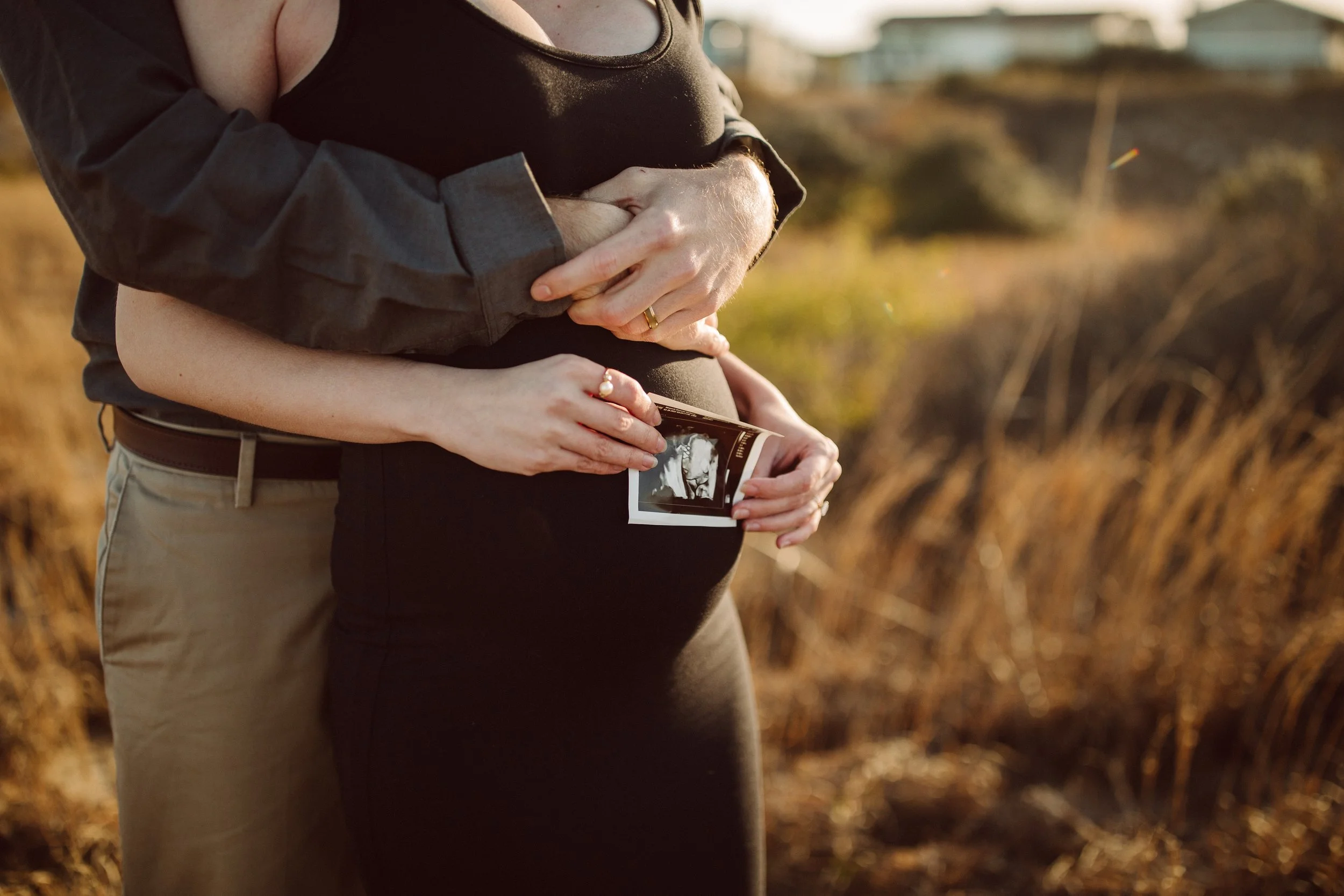 A pregnant woman is holding an ultrasound photo, with her partner's hands placed on her belly, standing outdoors in a field during sunset.