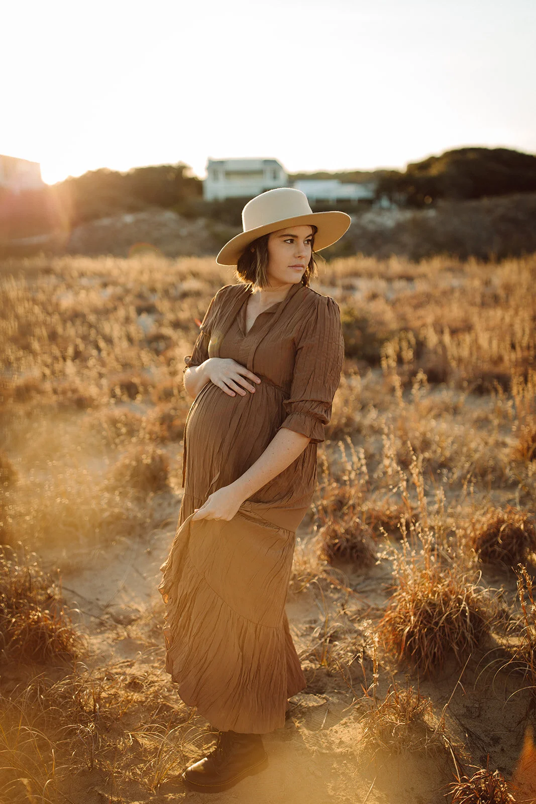 Pregnant woman in a brown dress and wide-brimmed hat standing in a dry grassy field during sunset.
