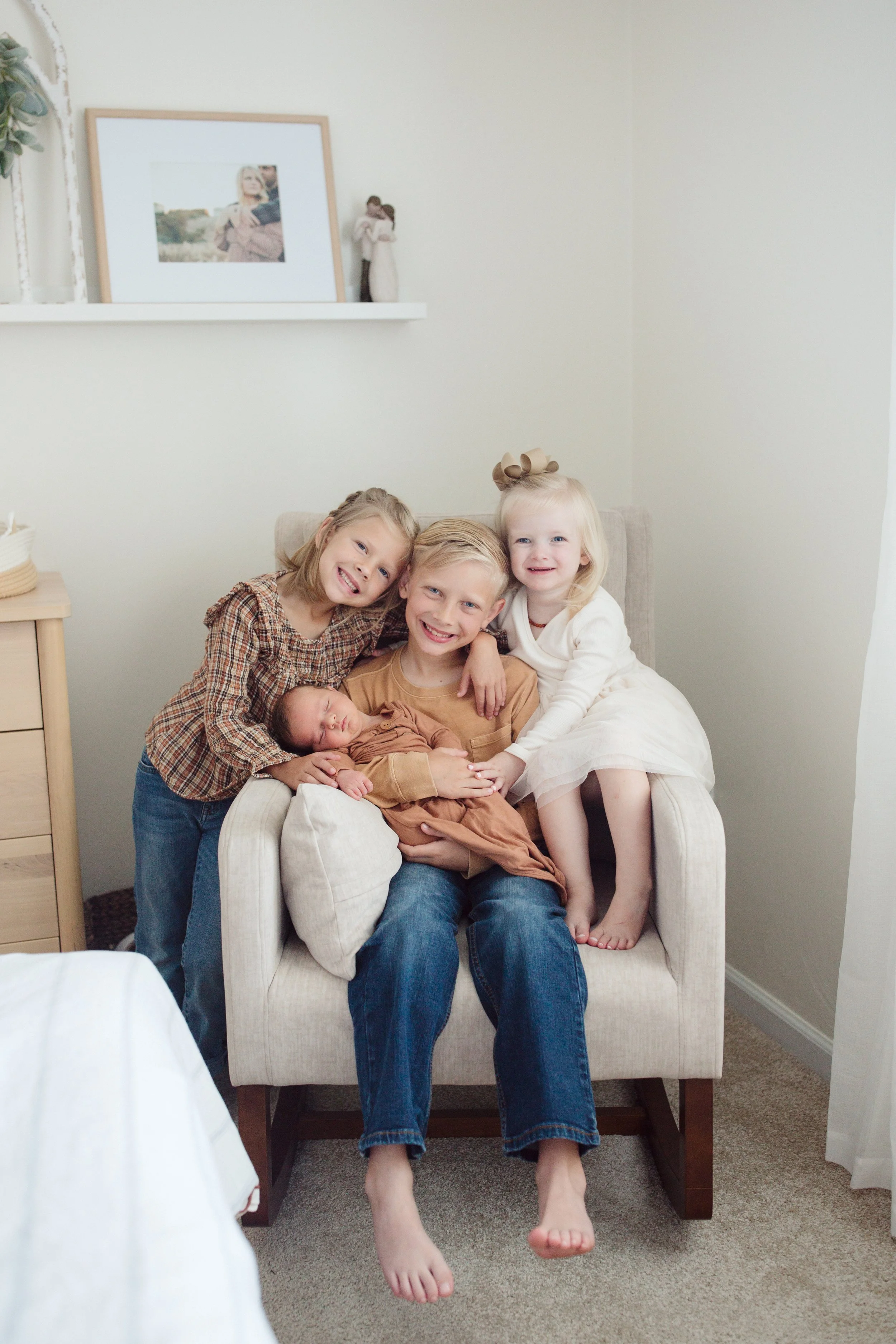 Four children sitting on a beige armchair inside a room, holding a newborn baby, with happy smiles. One girl has a large bow in her hair, another girl is wearing a plaid shirt, a boy in a light brown shirt, and a girl in a white dress. A framed pictu