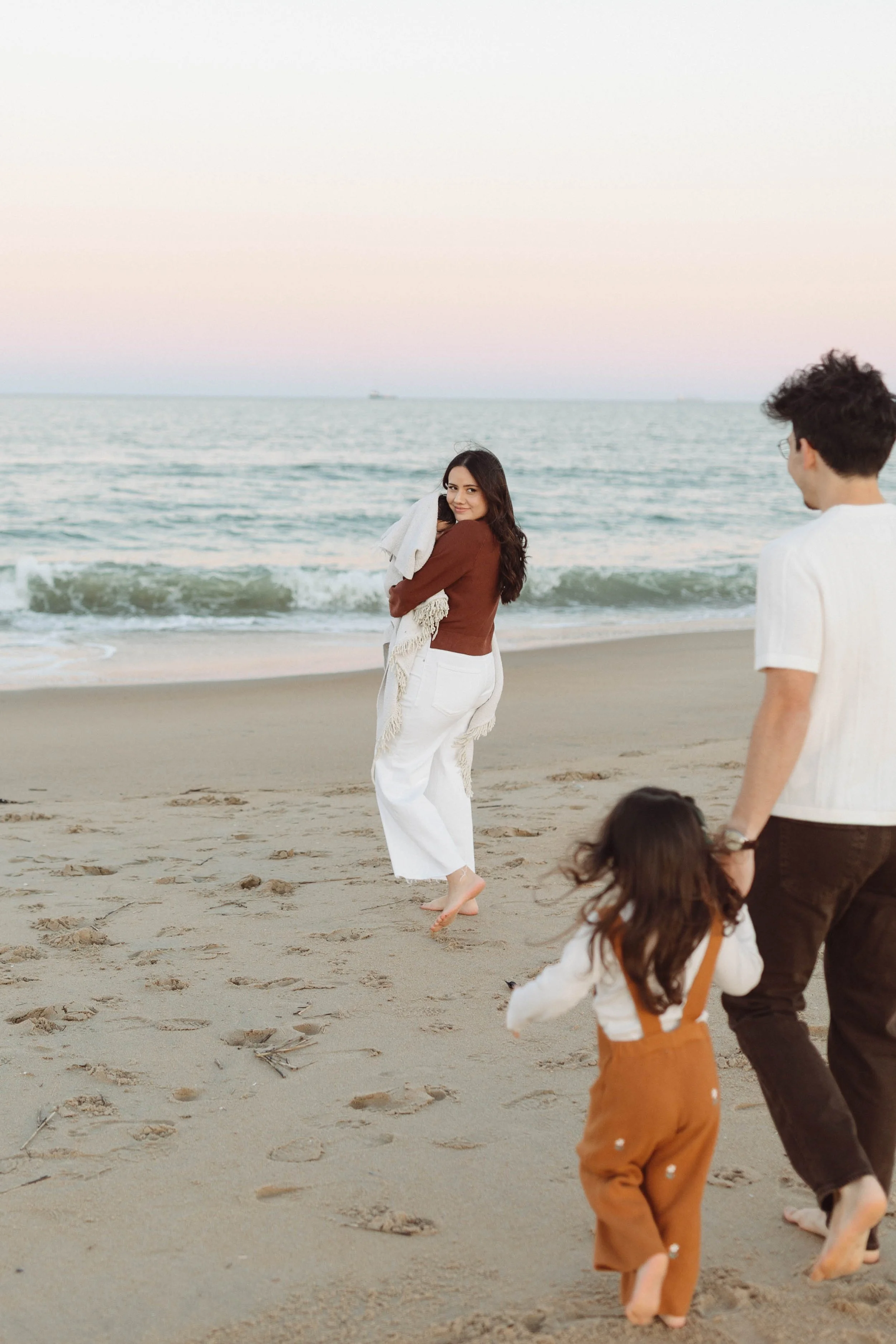 A woman and two children walking barefoot on the beach at sunset, with the woman holding a blanket and smiling at the camera.