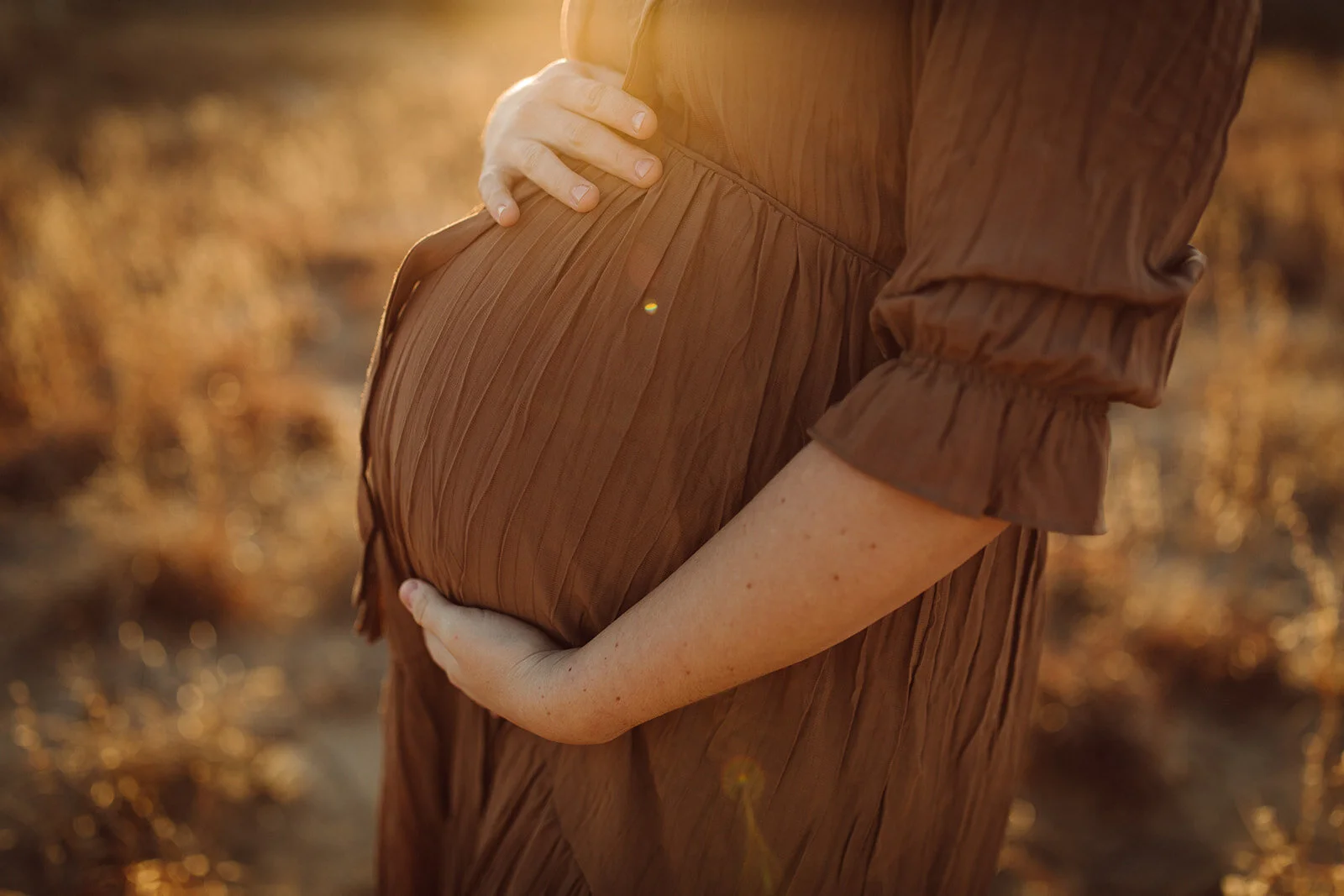 A pregnant woman wearing a brown dress gently holds her belly with both hands in a field during sunset.