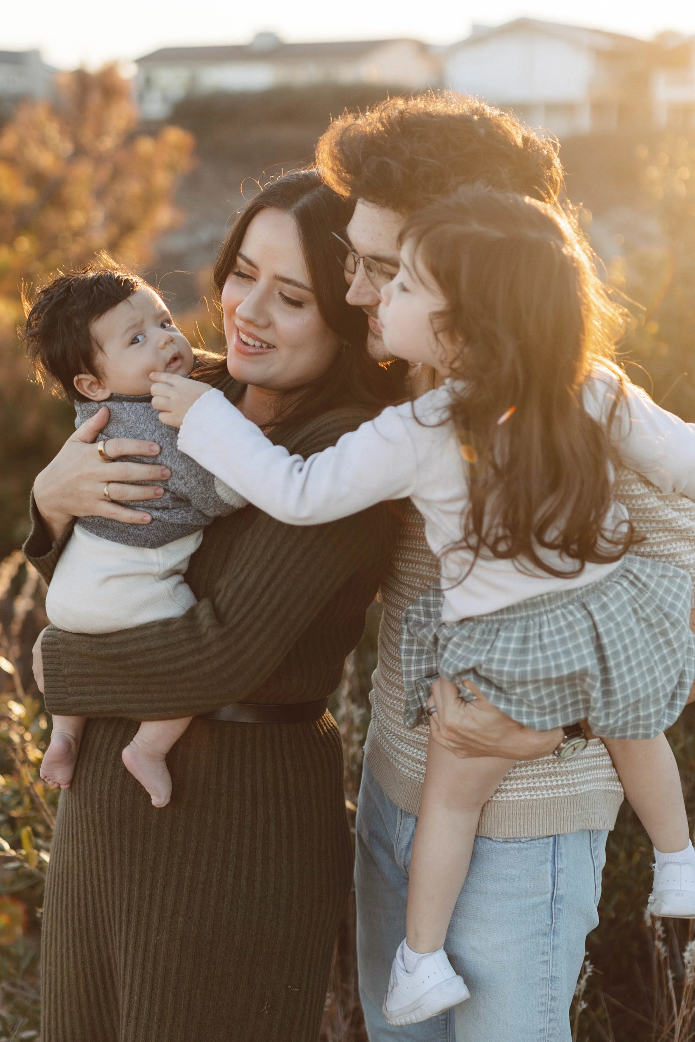 A family of four, including a mother, father, and two young children, enjoying a moment together outdoors during golden hour.