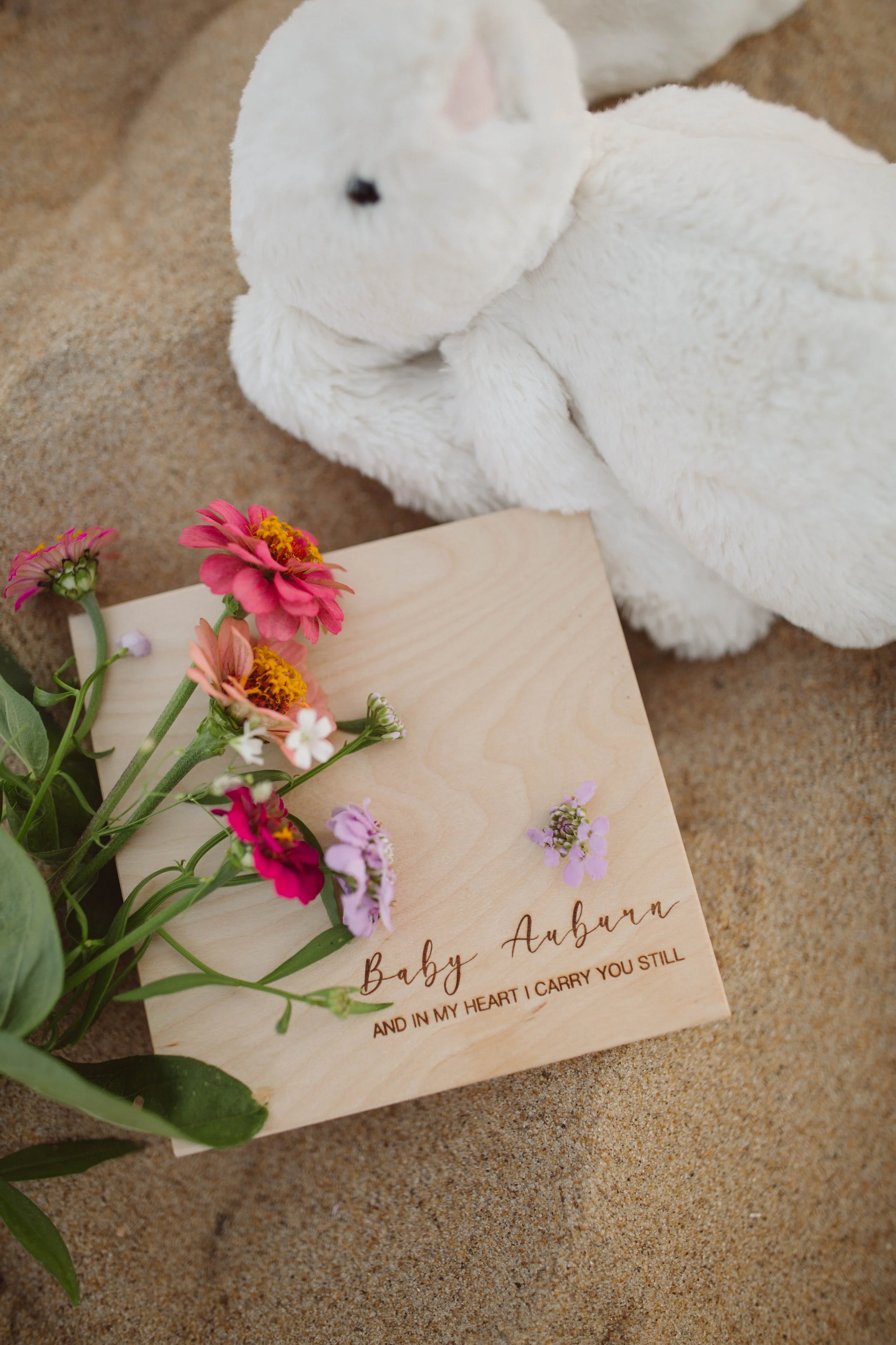 A plush white bunny stuffed animal, a wooden sign with the text 'Baby Auburn and in my heart I carry you still', and a small bouquet of pink and purple flowers on a sandy surface.