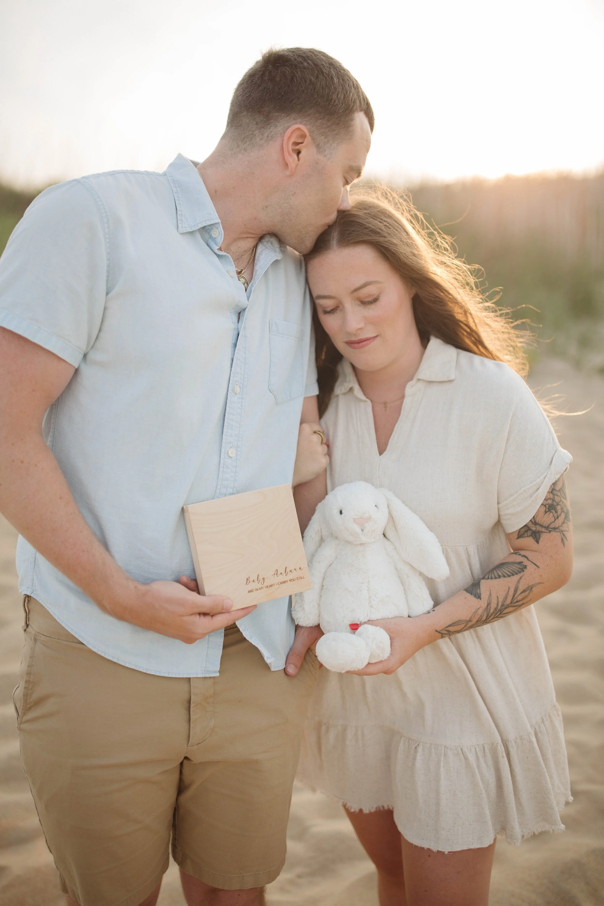 A couple stands close together on a beach, the man kissing the woman on her forehead. The woman is holding a plush white bunny toy and a wooden box with the inscription 'Baby Aurora'.