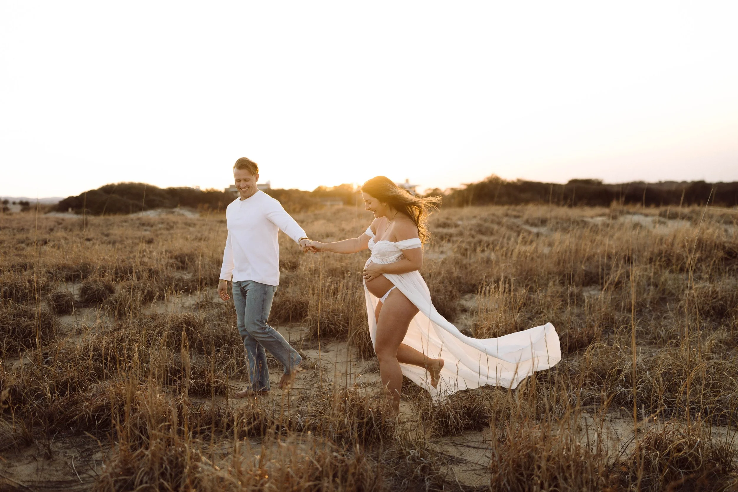 couple holds hands and smiles as they walk over golden dunes at virginia beach oceanfront while woman cups pregnant belly and wears a white sheer flowing gown