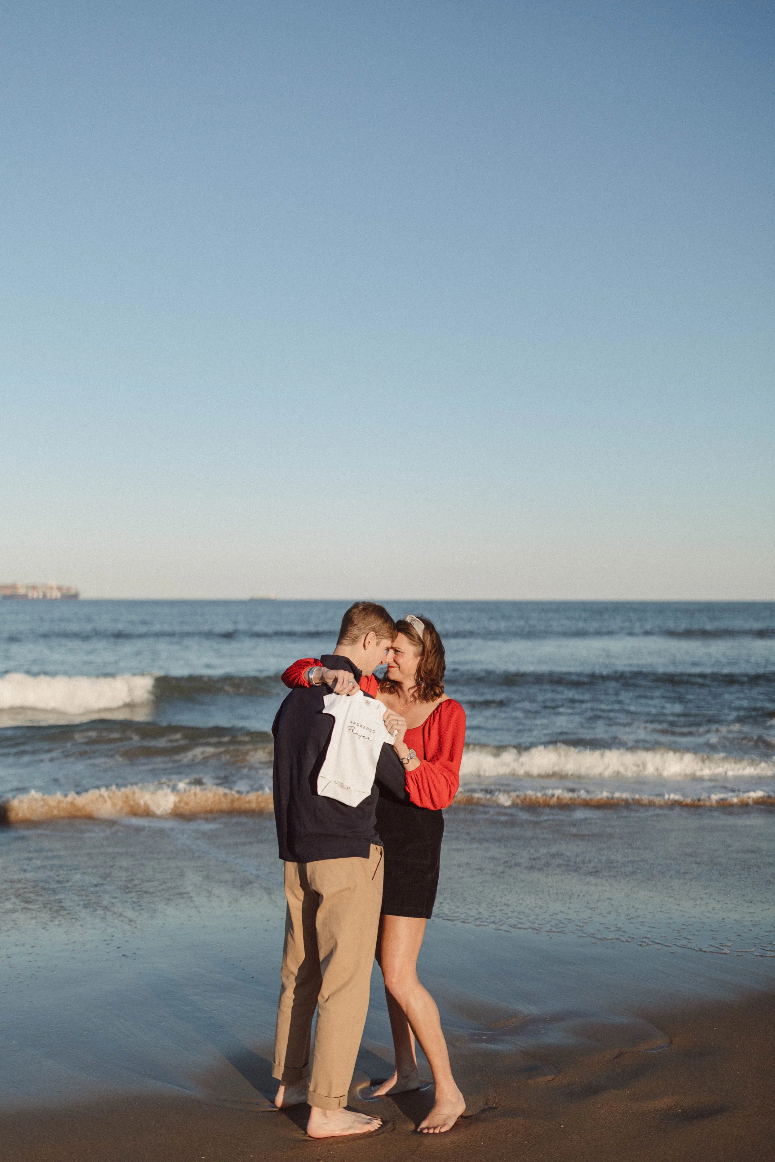 virginia beach military couple stands in front of ocean holding a onesie for maternity pictures