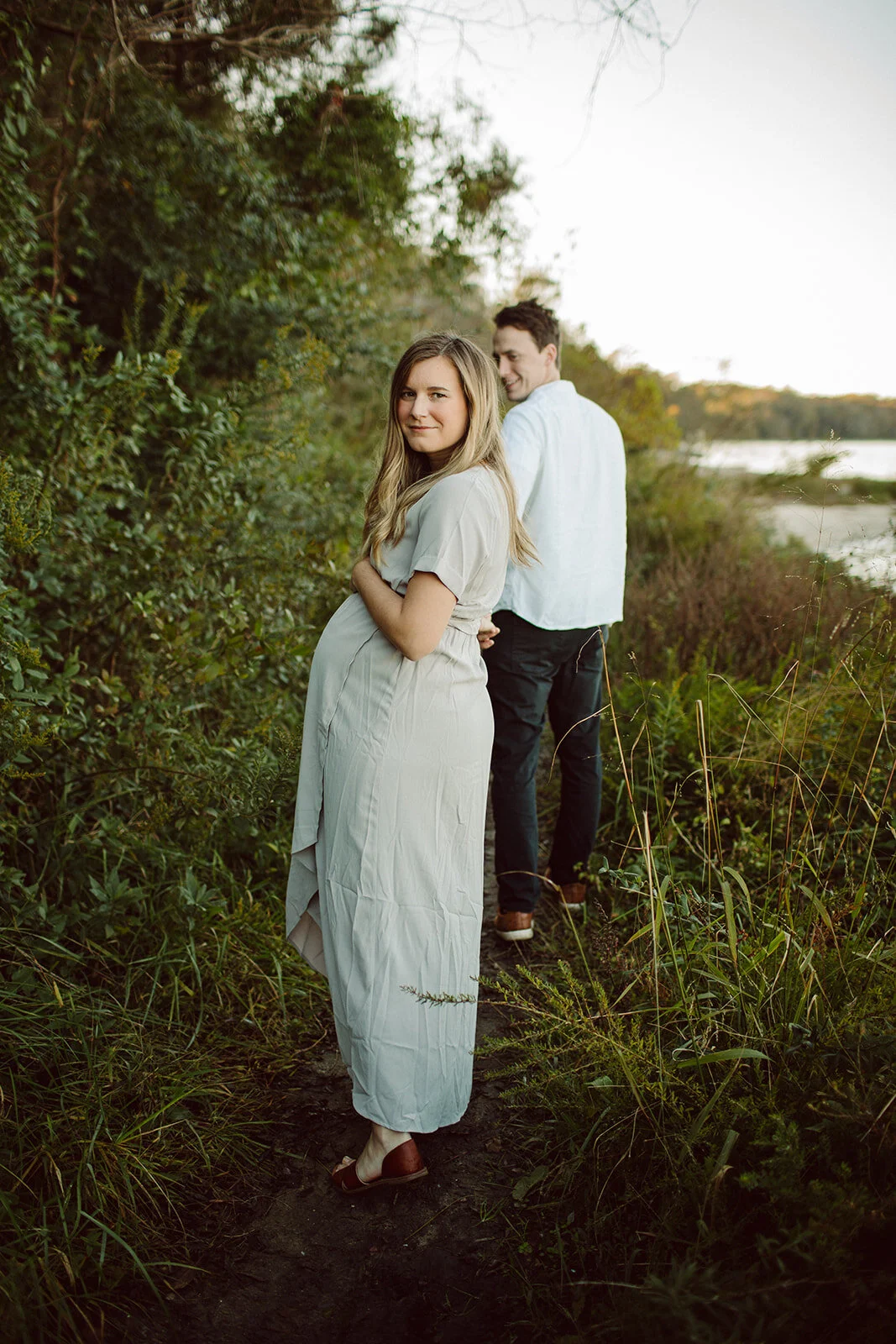 A couple standing on a trail near a body of water, surrounded by greenery. The woman in the foreground wears a light-colored dress and looks at the camera, while the man behind her looks away towards the water.