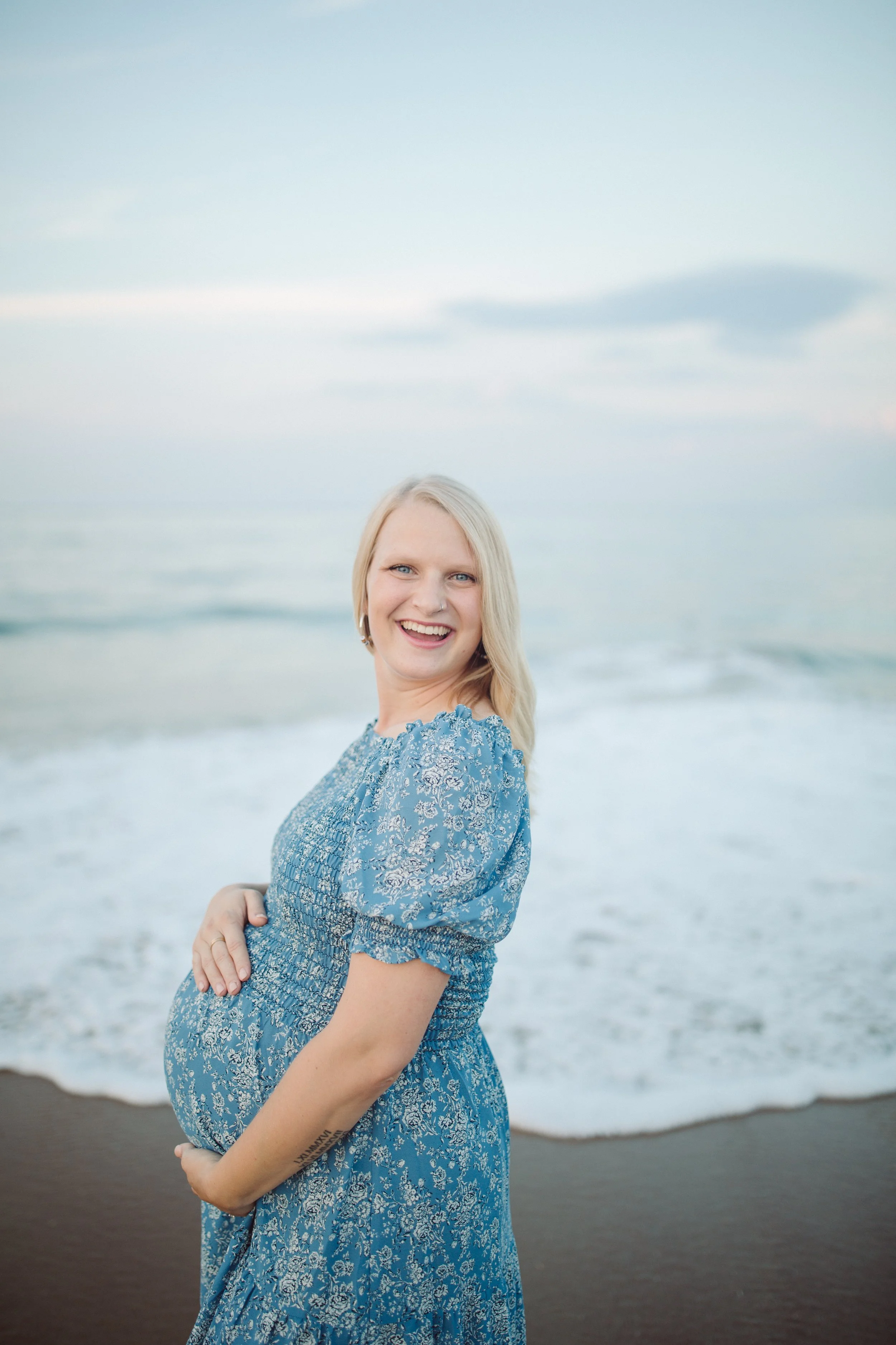 A smiling pregnant woman in a blue floral dress standing on a beach with the ocean and sky in the background.
