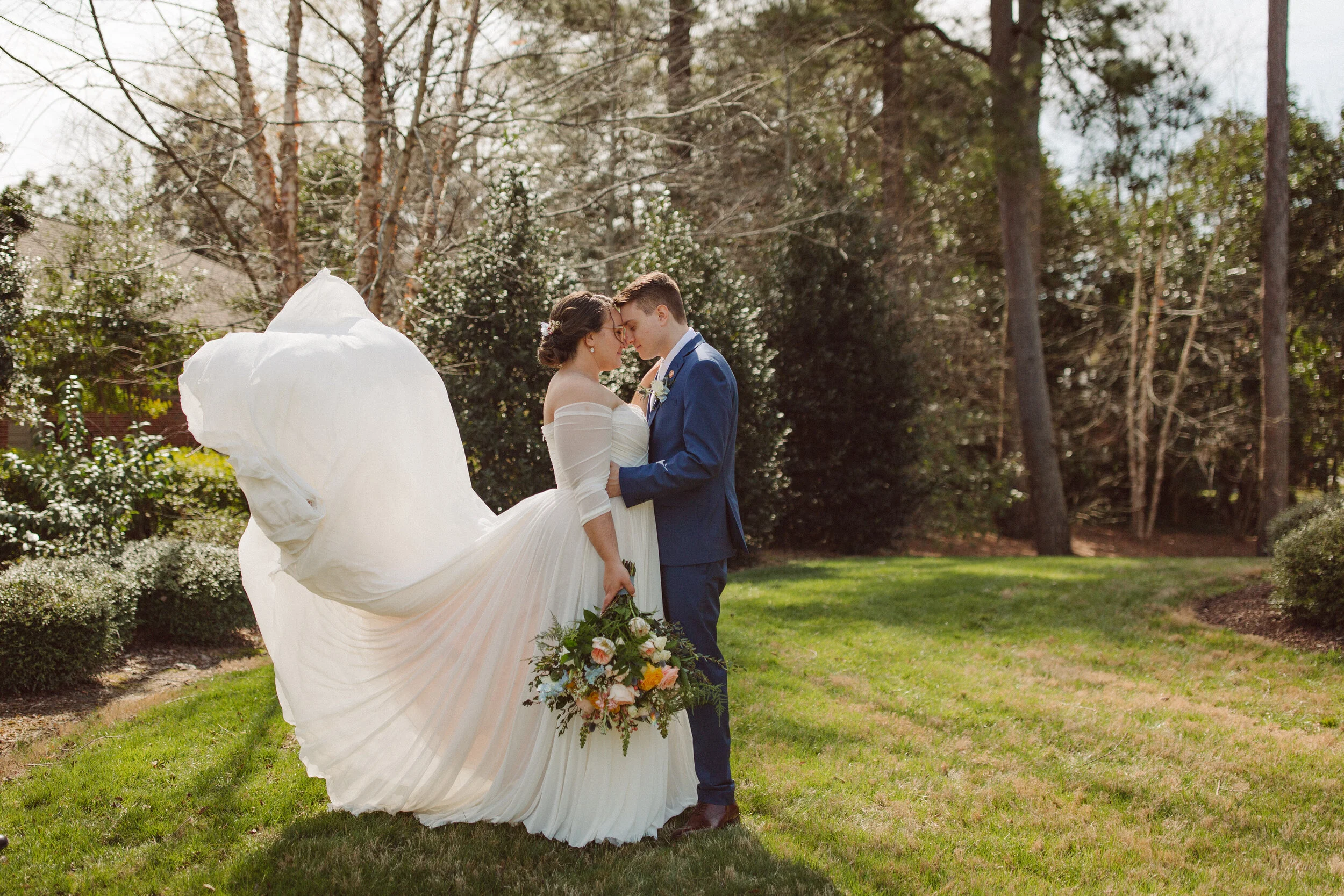 A bride in a flowing white wedding dress holding a bouquet and a groom in a blue suit in an outdoor garden, standing close with foreheads touching. Tall trees and bushes surround them.