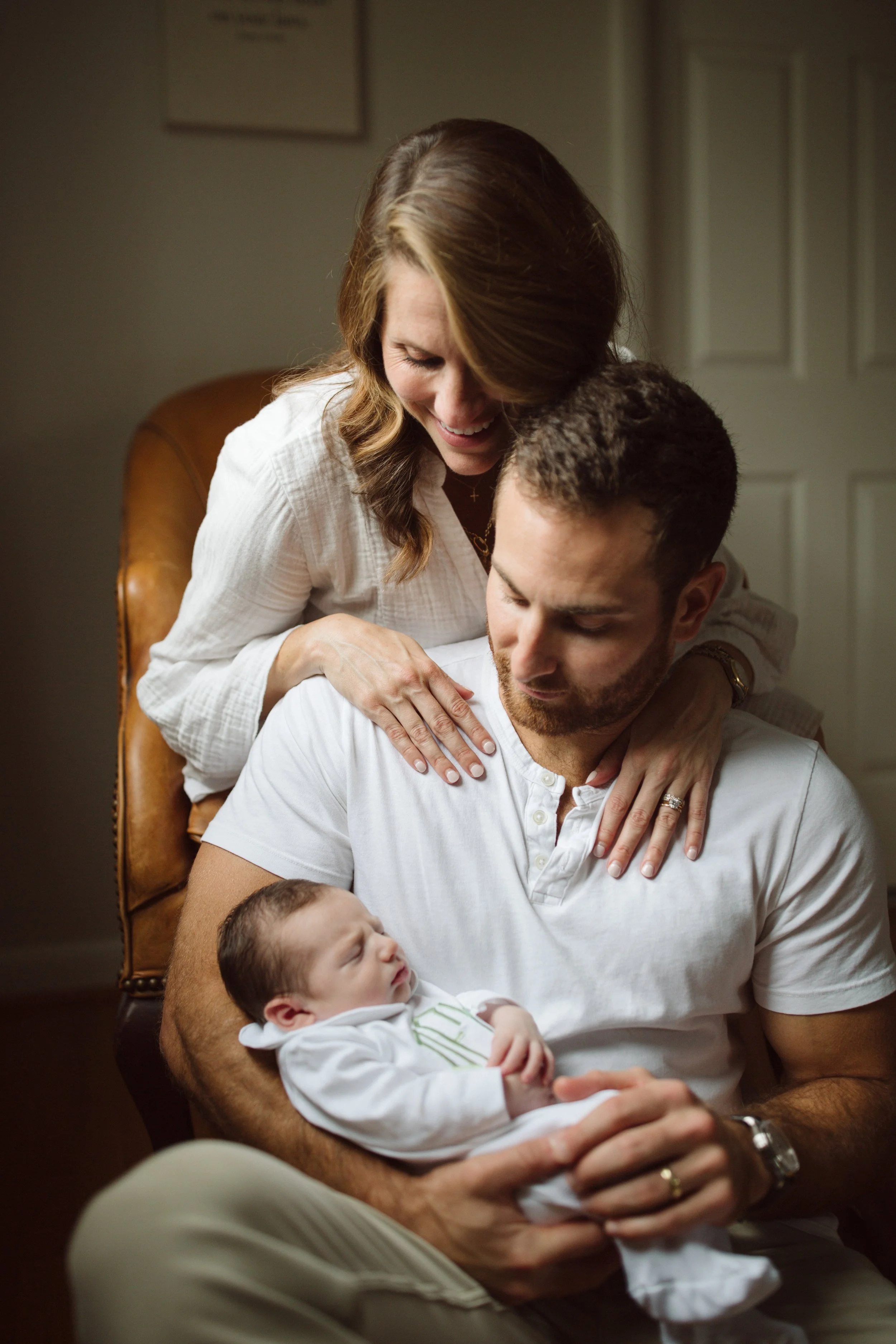 A happy woman leaning over a man holding a sleeping baby, all sitting in a cozy indoor setting.