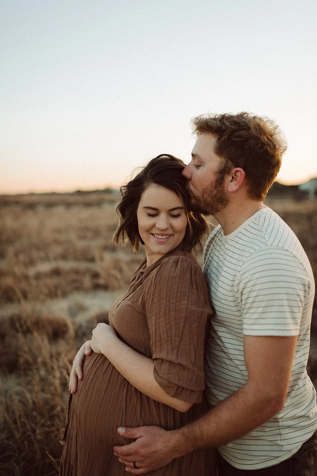 A couple, with the man kissing the woman's forehead, stands in a field during sunset, she is visibly pregnant and smiling.