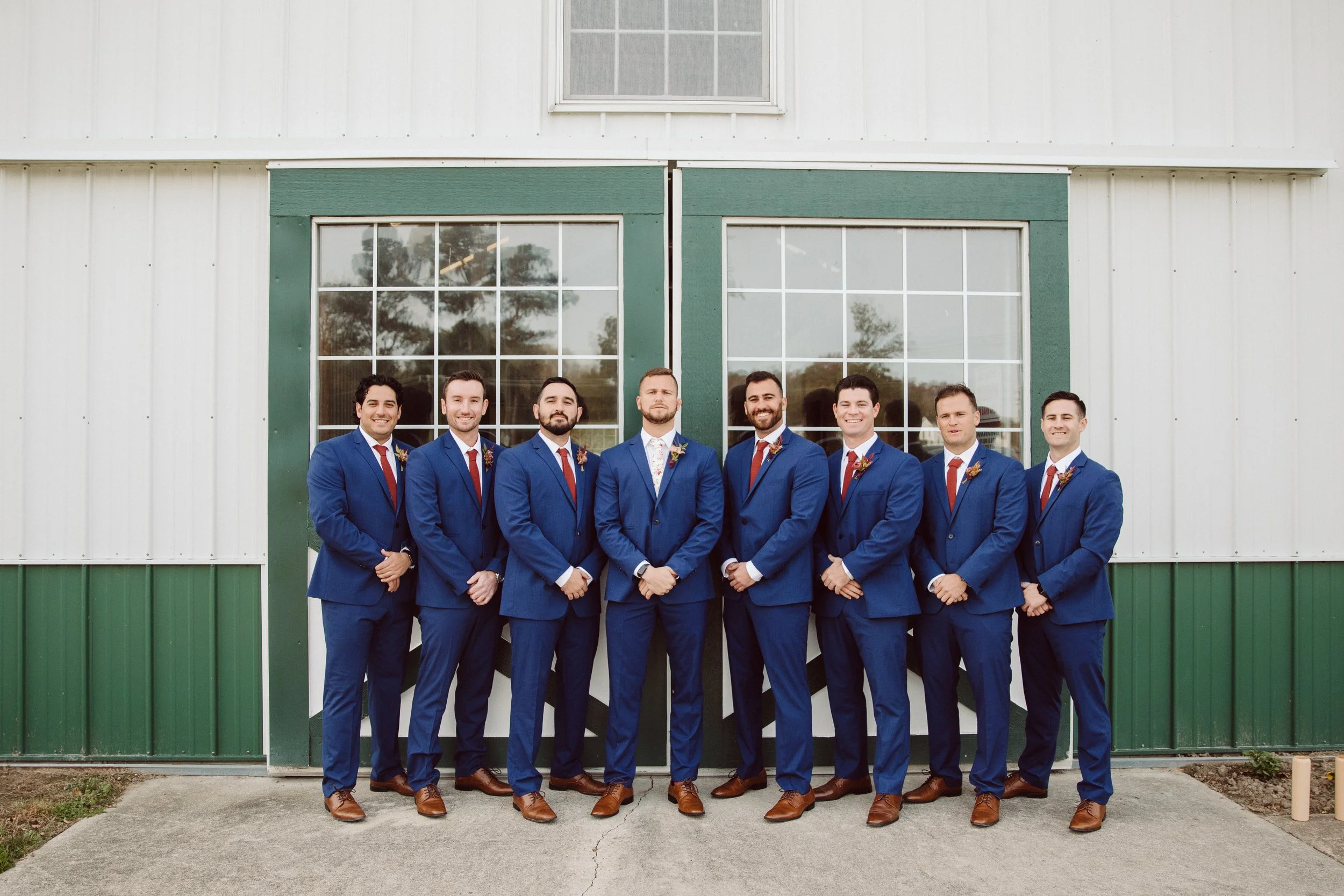 Group of eight men in blue suits and red ties standing in front of a green and white barn, smiling.