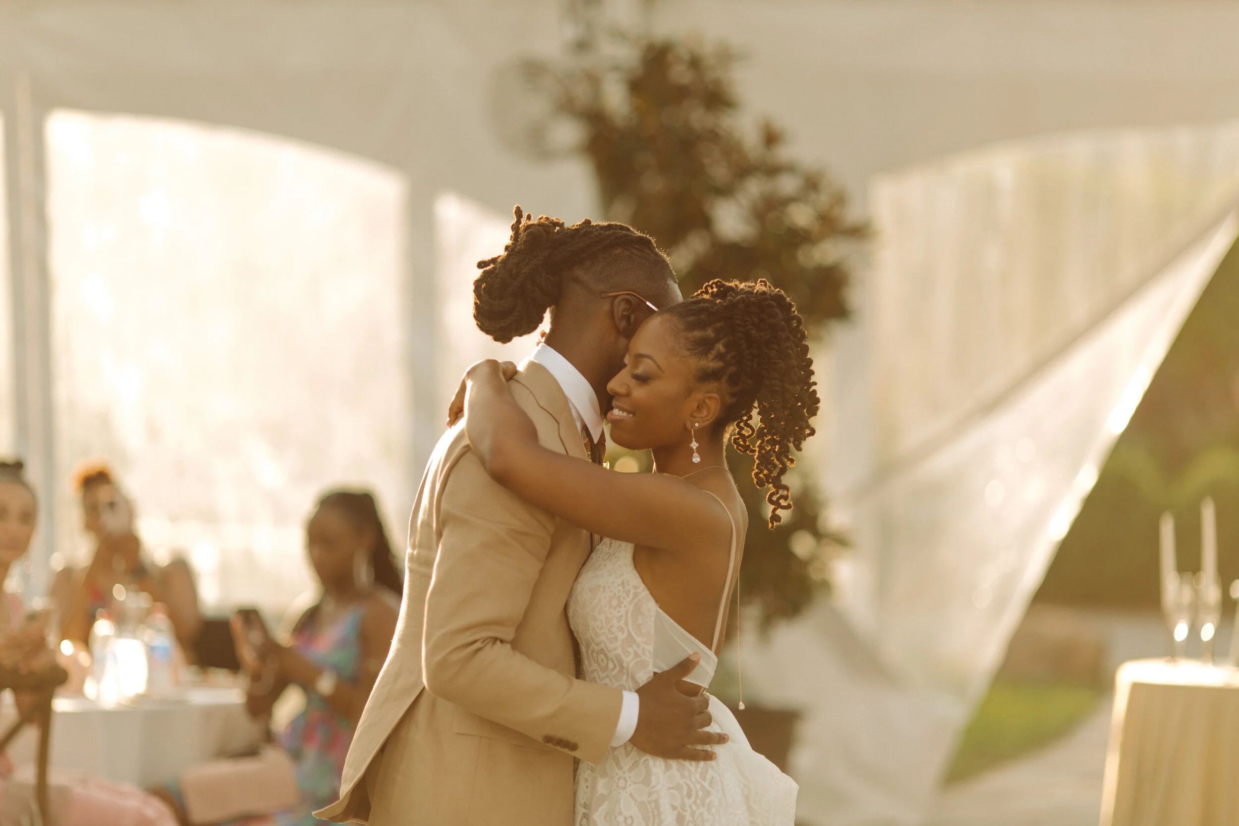 A bride and groom share a dance at their wedding reception, embracing tenderly and smiling in a warmly lit, elegant venue.