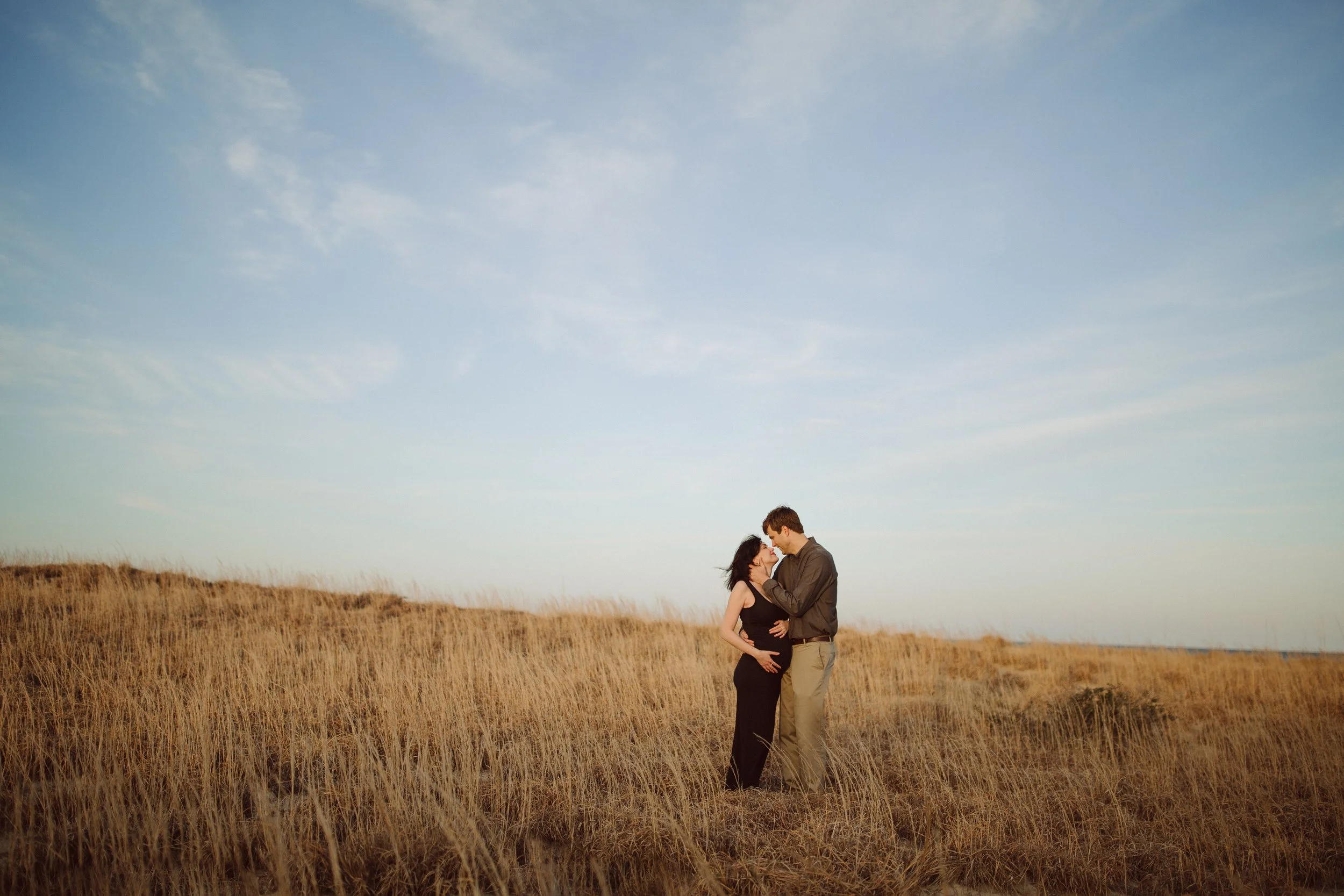 A couple standing close together in a grassy field, holding each other affectionately under a blue sky with scattered clouds.