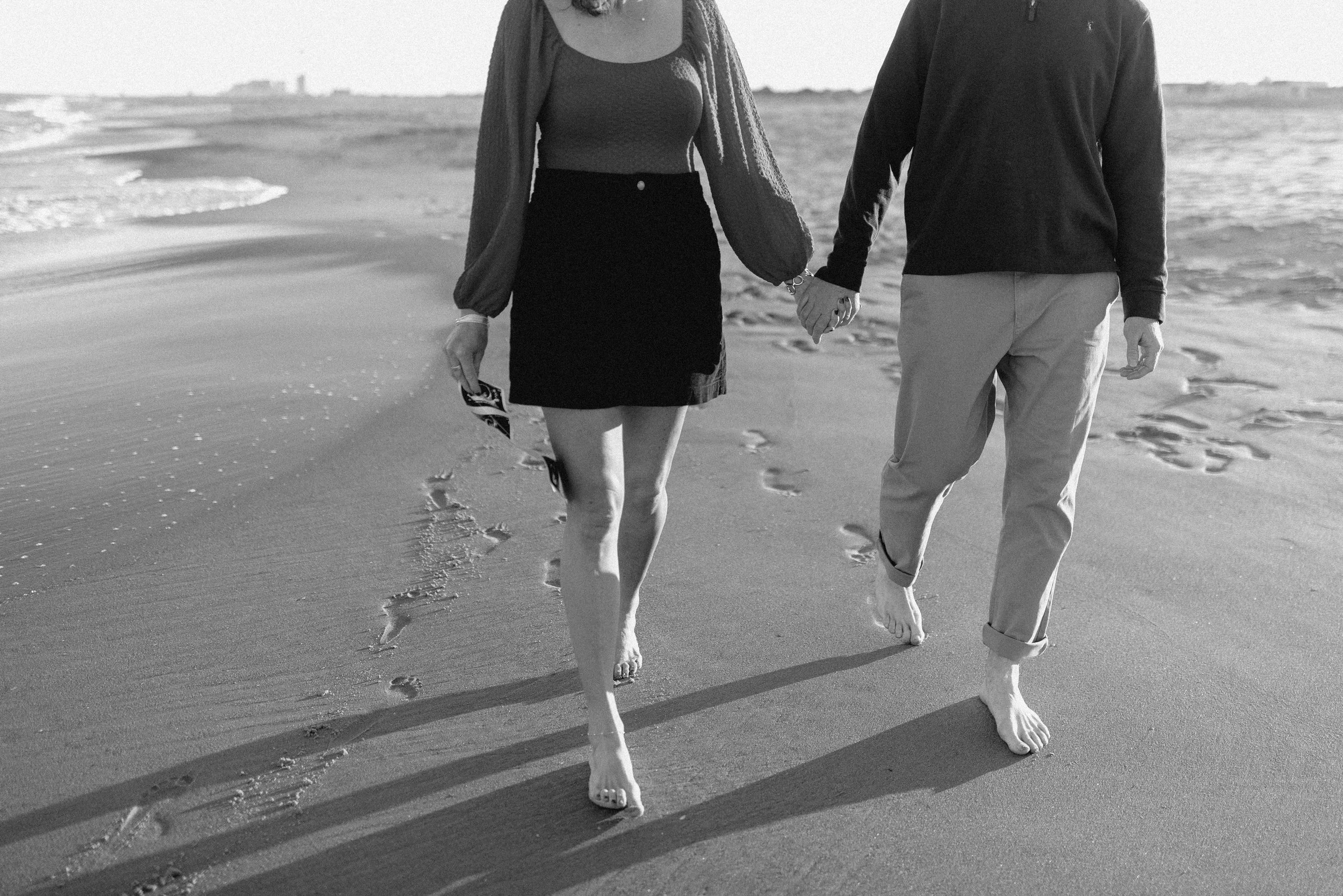 black and white picture of couple walking down beach holding ultrasound pictures with sand and virginia beach oceanfront behind them.