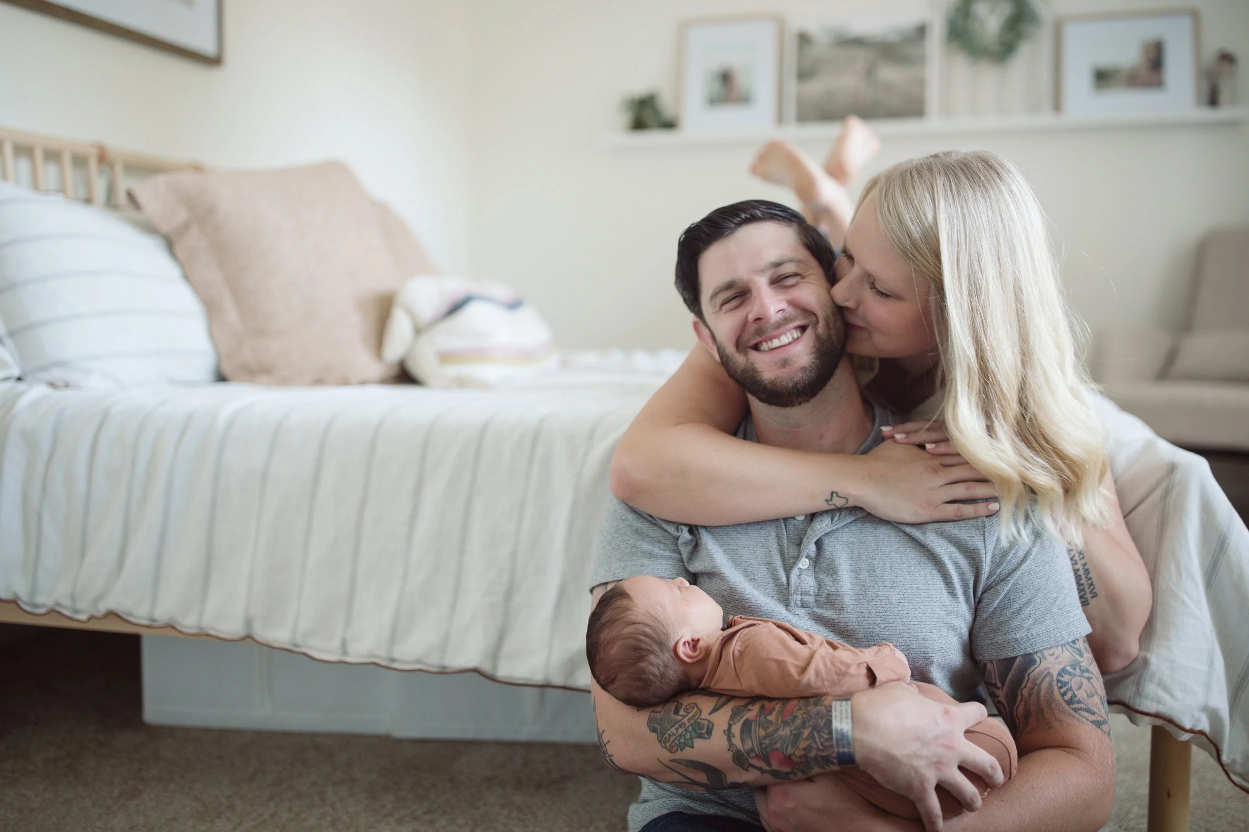 A happy family with a man, woman, and baby in a cozy bedroom, the woman kissing the man on the cheek, the man holding the baby, all smiling.