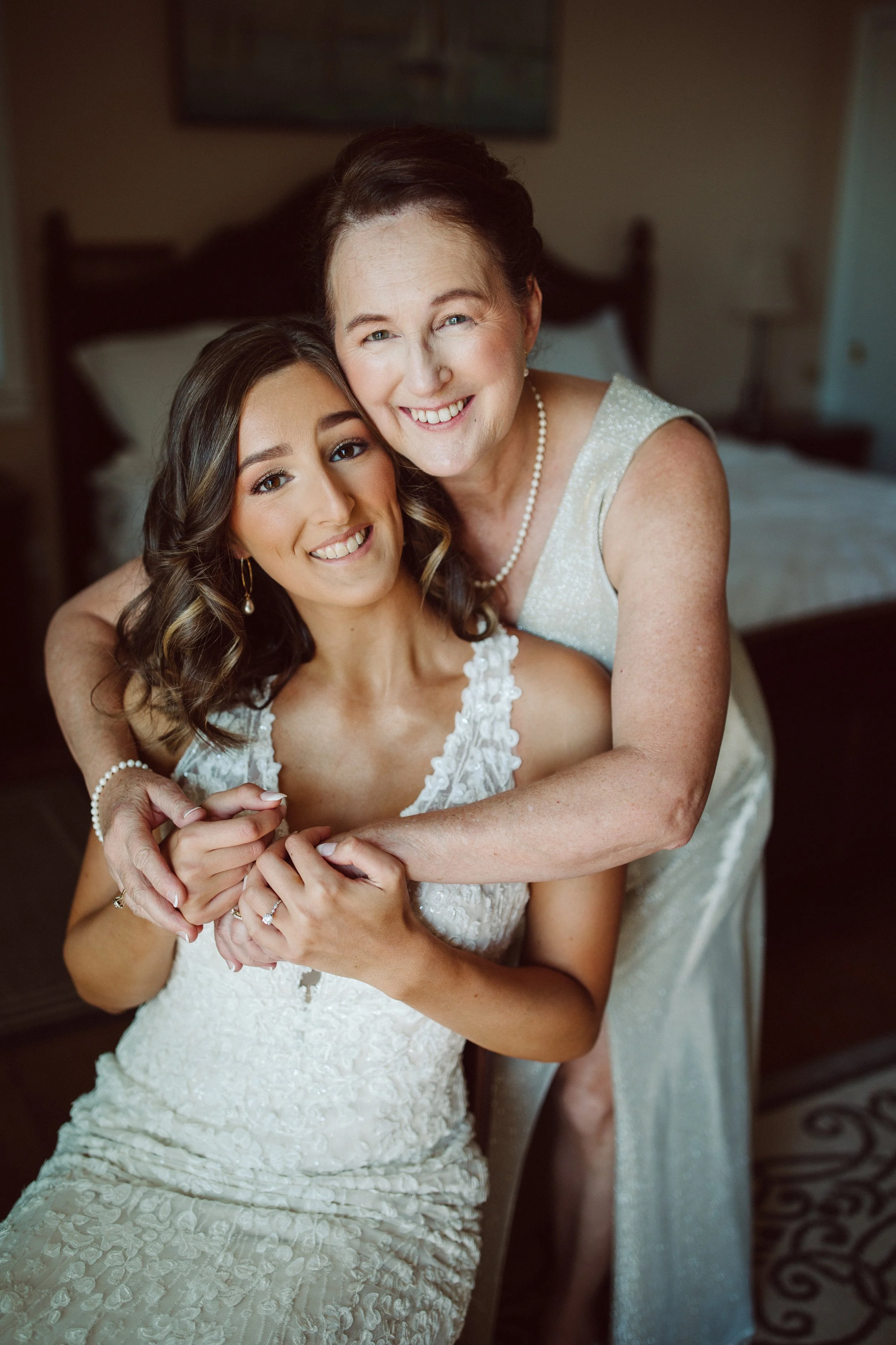 A young woman in a wedding dress and a smiling older woman in a silver dress embrace in a warmly lit bedroom.