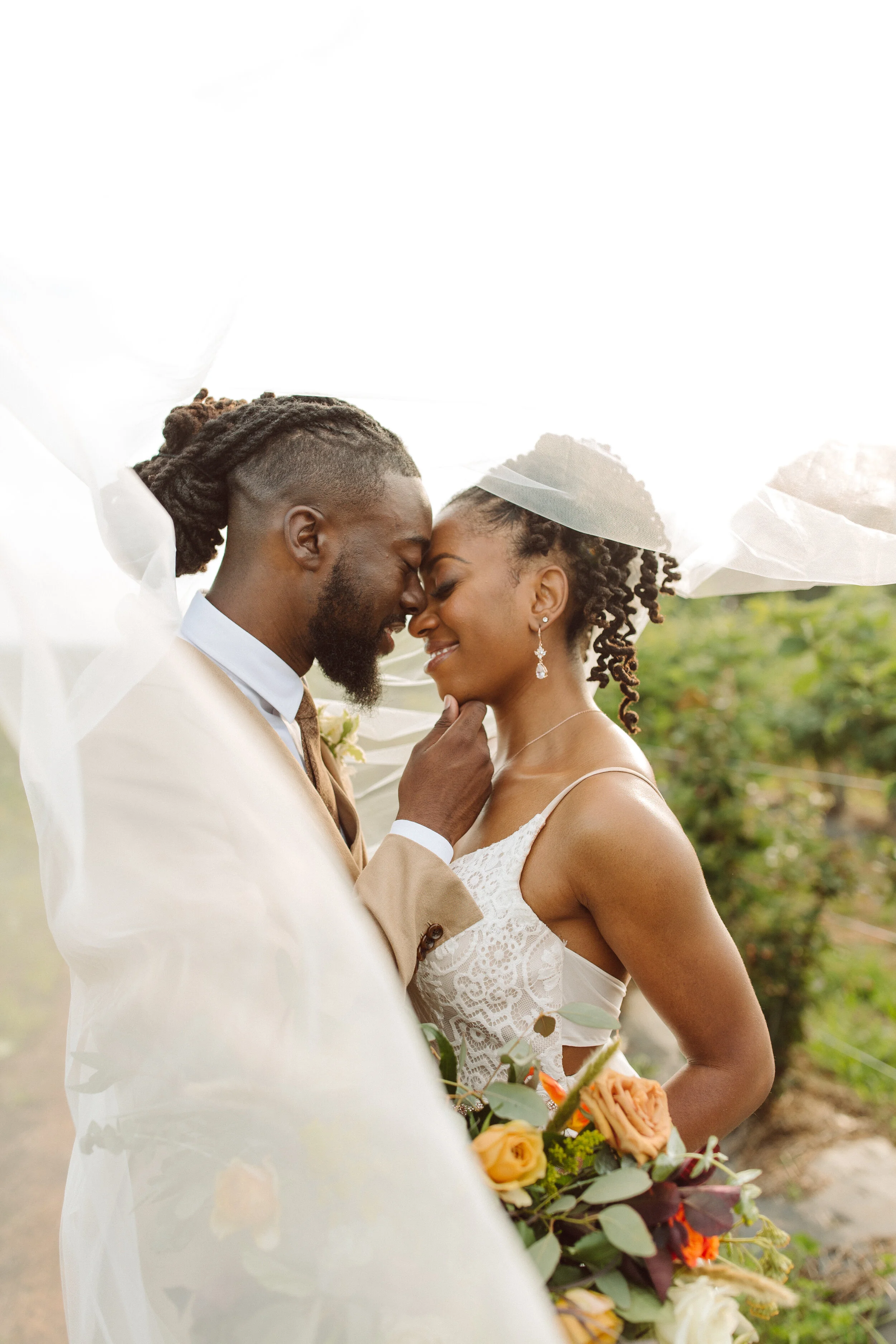 A newlywed couple sharing a loving moment under a white veil outdoors, with the groom in a beige suit and the bride in a lace wedding dress holding a bouquet of yellow and orange roses.