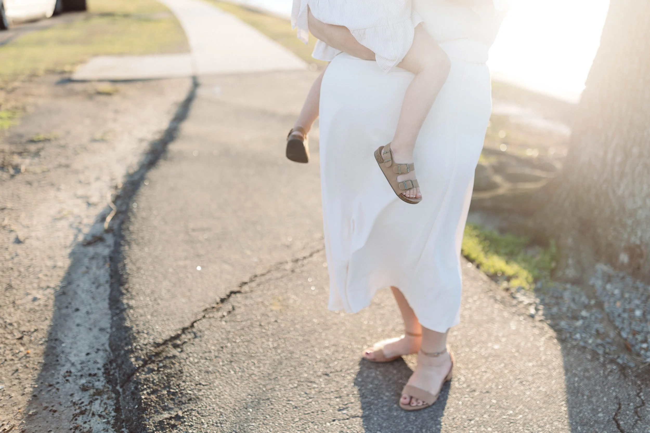 mom in white dress holds little girl in white dress over her pregnant belly with sun shining behind them.