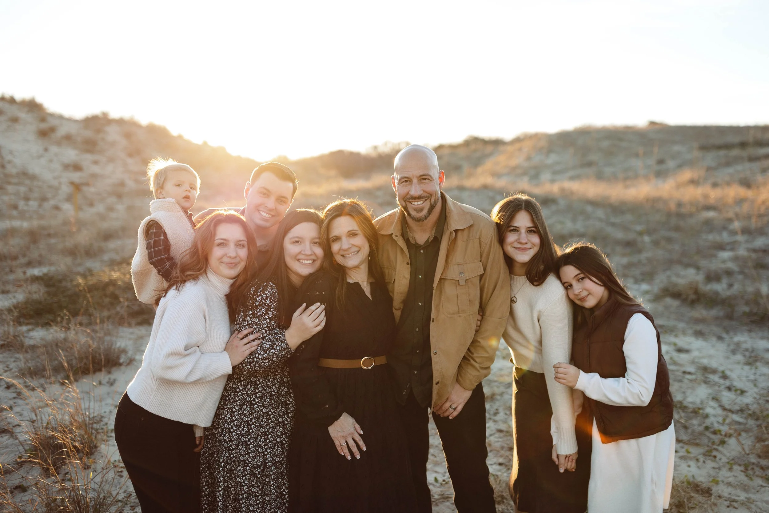 family stands hugging and smiling at the camera wearing coordinating attire of blacks, neutrals, and whites. They are standing in between sand dunes as the sun shines behind them near Back Bay Wildlife Refuge