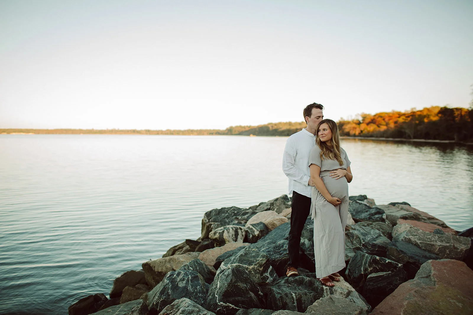 A pregnant woman and her partner standing on rocks by a lake during sunset, with trees in fall colors in the background.