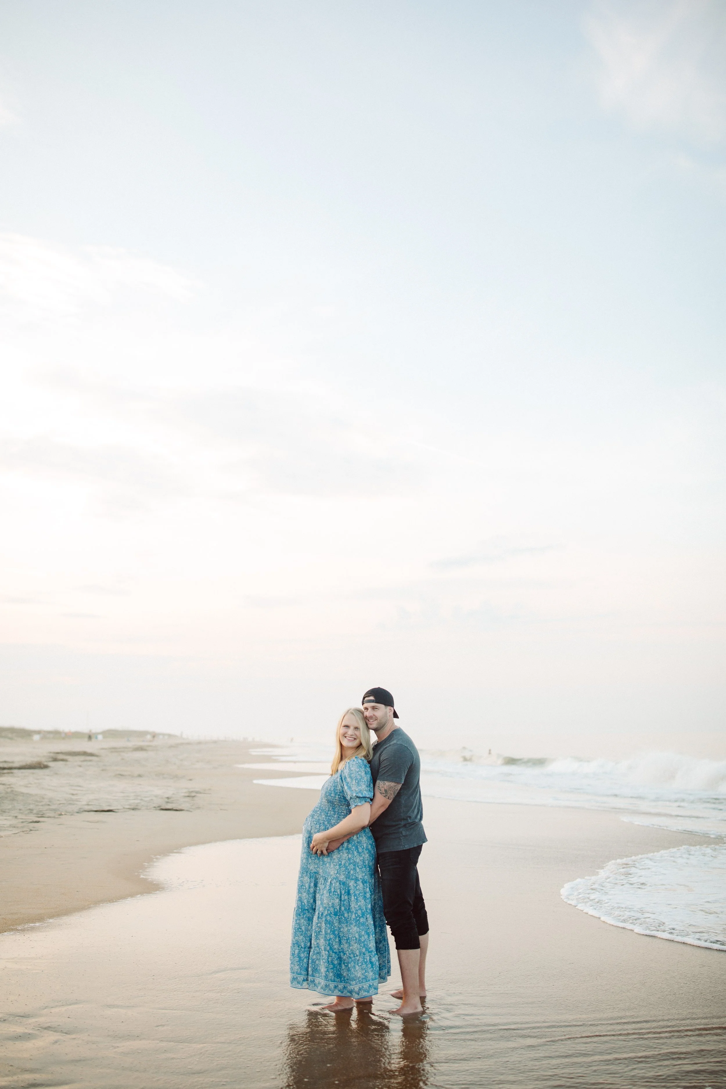 A smiling pregnant woman in a blue dress standing in the shallow surf with a man in a dark t-shirt and shorts, both barefoot, on a beach during sunset.