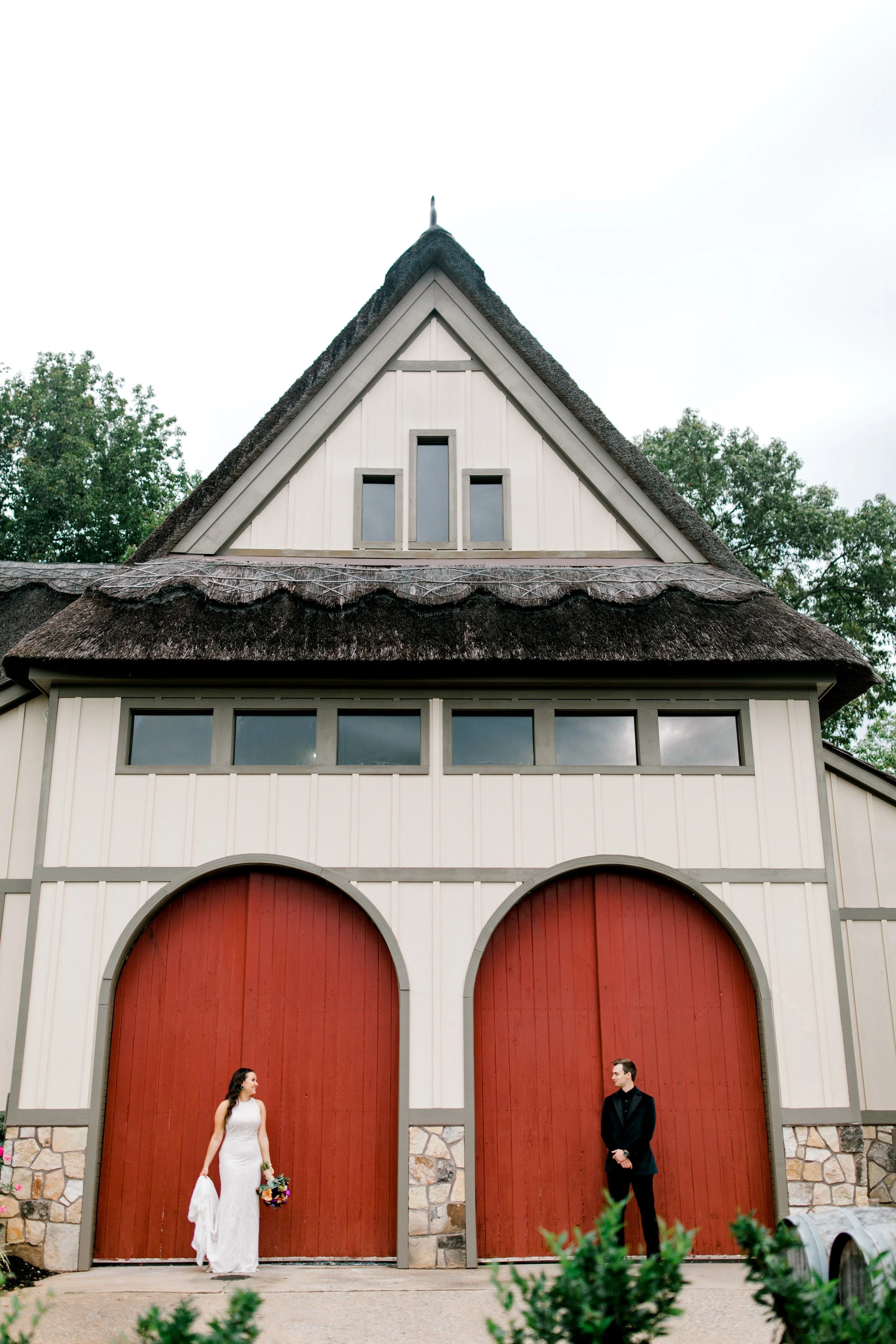 A bride and groom stand outside a large barn with red doors, the bride holding a bouquet and wearing a white dress, the groom dressed in black, in front of the barn's front doors.