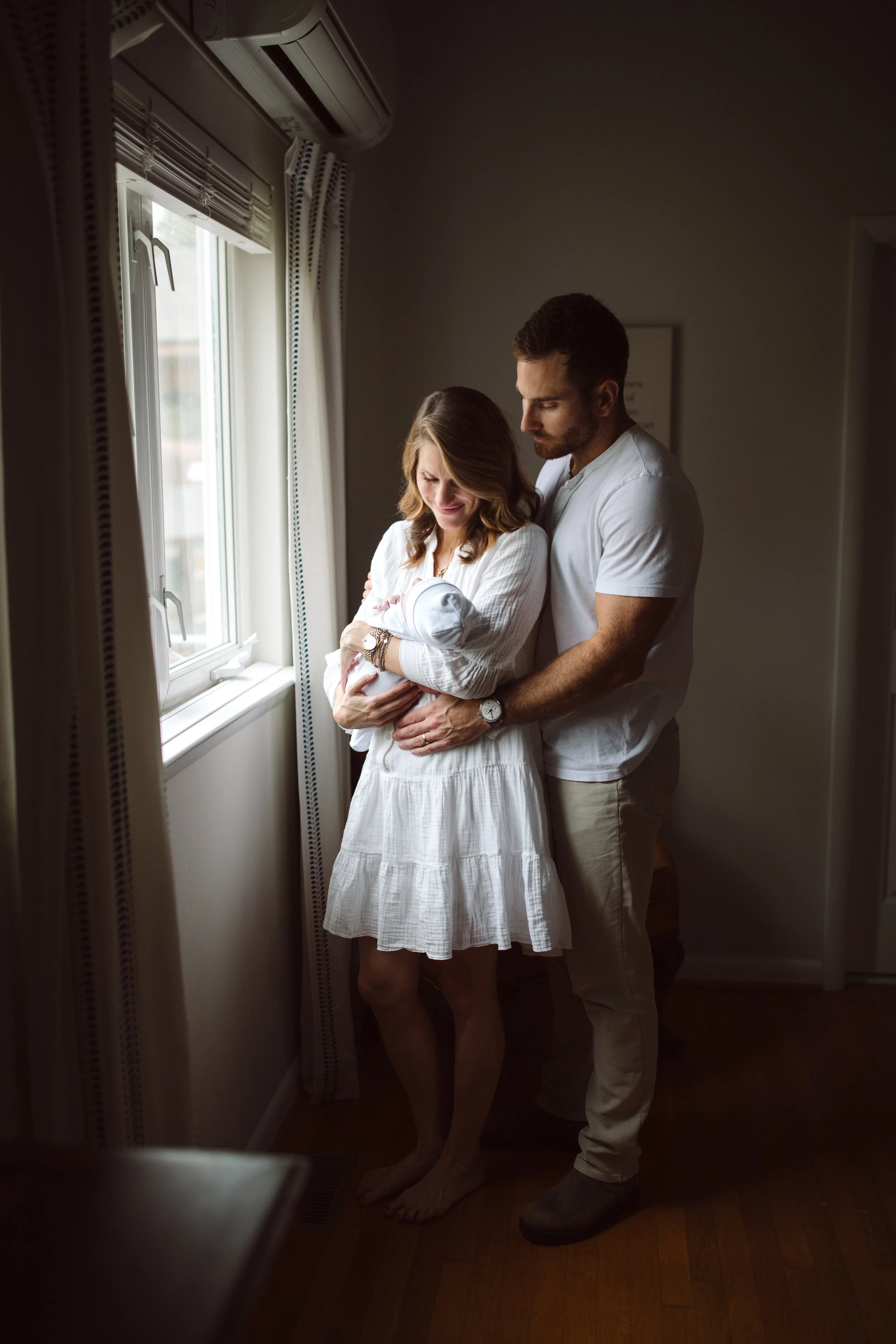 A couple holding a newborn baby by the window in a softly lit room, the woman is smiling and the man is gently holding her from behind.