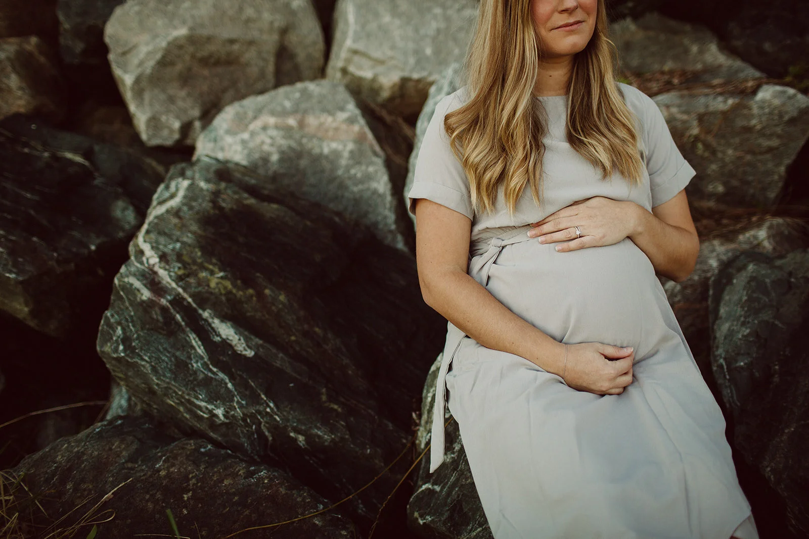 A pregnant woman sitting on rocks, gently holding her belly with one hand and her stomach with the other, outdoors.