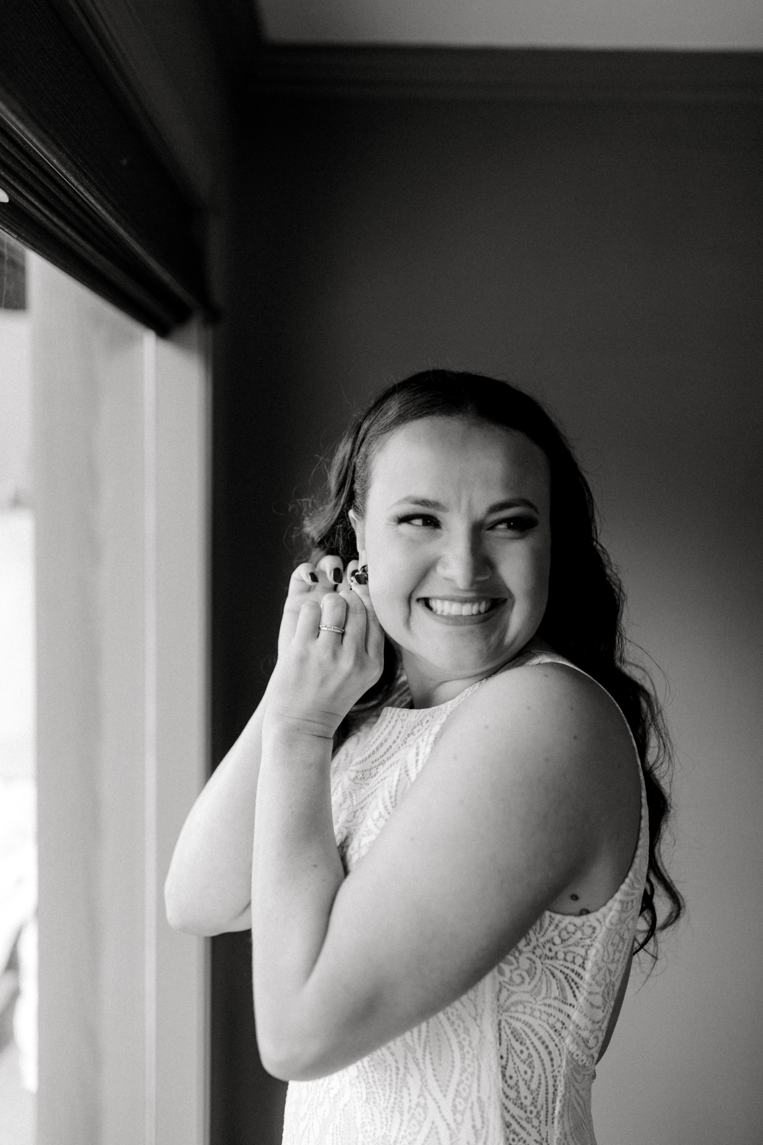 A smiling woman with long curly hair putting on earrings near a window, wearing a sleeveless lace dress.