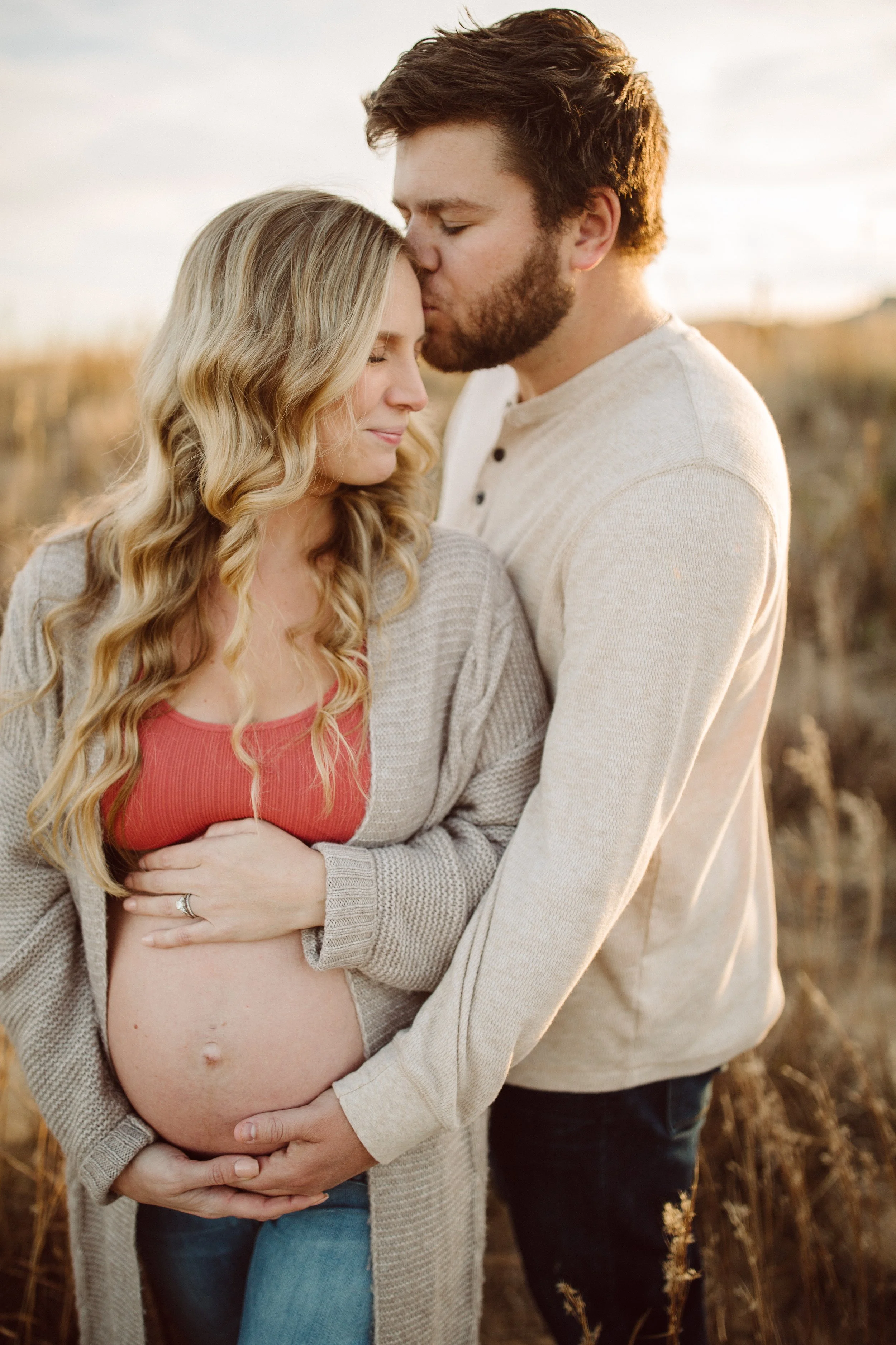 A pregnant woman and a man sharing a tender moment outdoors at sunset, with the man kissing the woman's forehead as she cradles her baby bump.