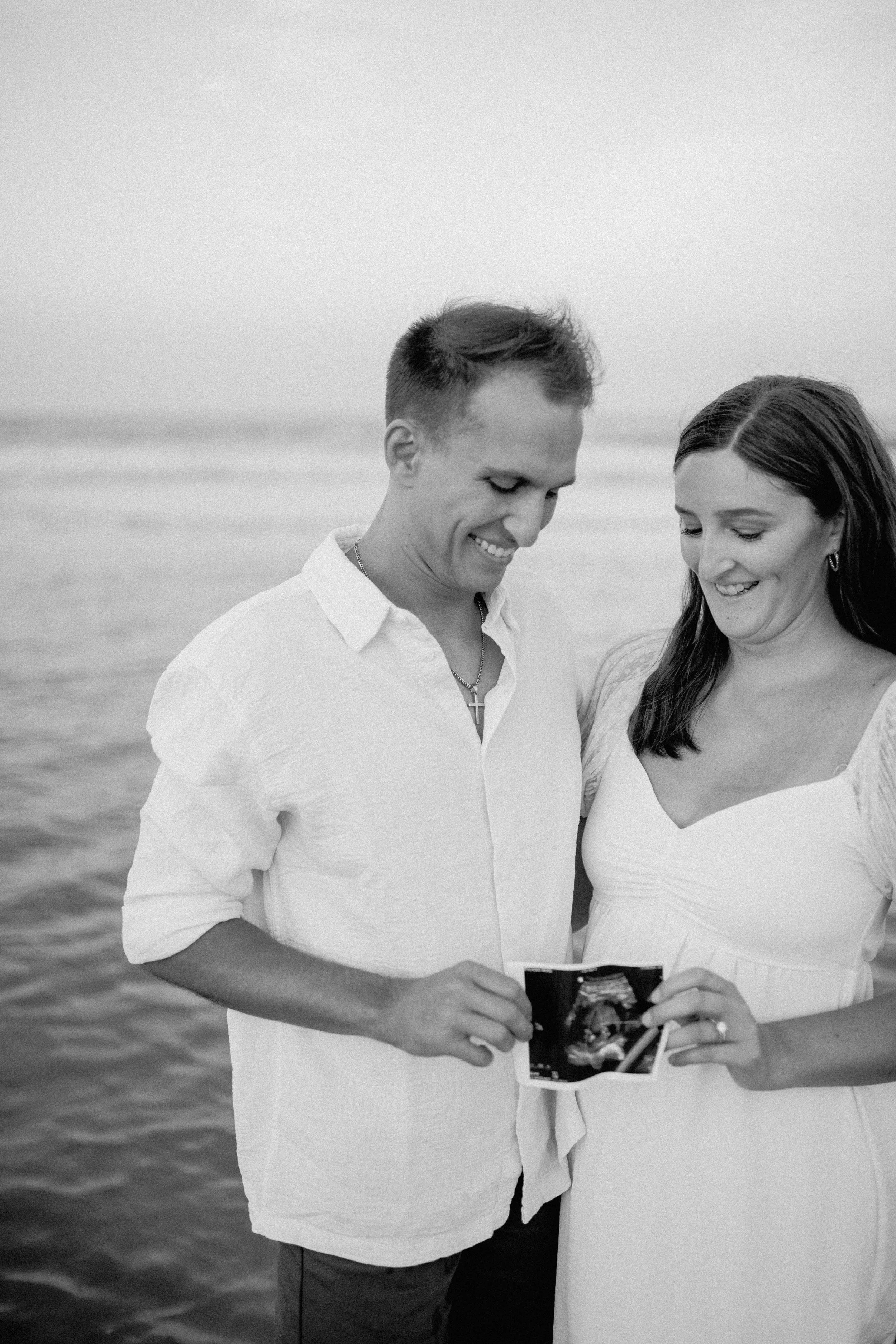 A couple standing by the water, looking at an ultrasound photo together, smiling, at the beach.