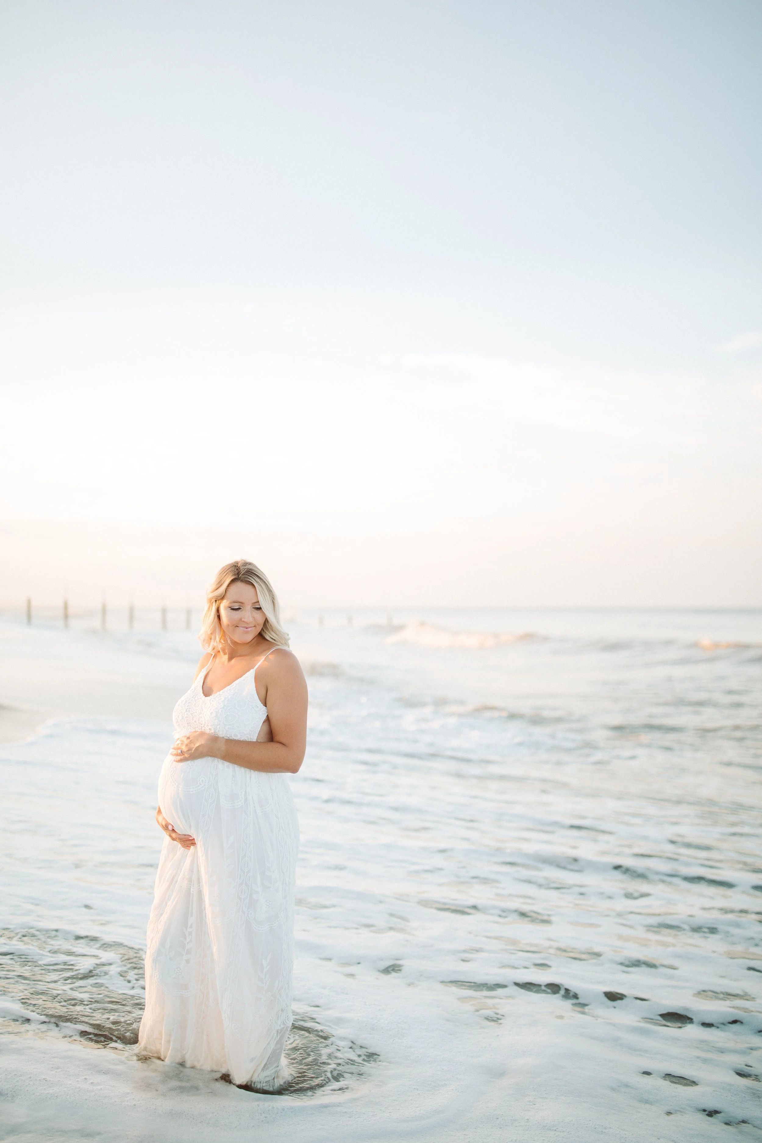 Pregnant woman in a white dress standing in the ocean water at the beach during sunset.